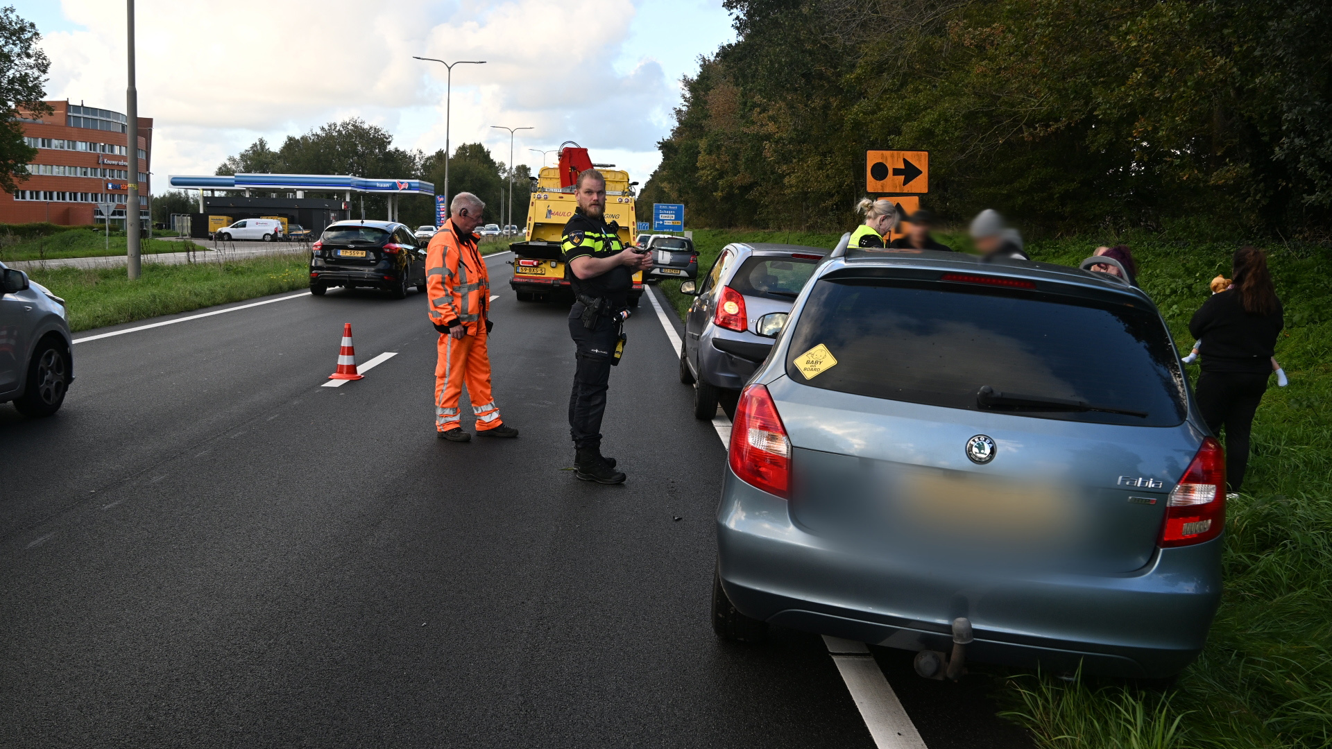 Politieagent en wegwerker regelen verkeer bij een stilstaande auto langs de weg, met pionnen en pechhulpwagen