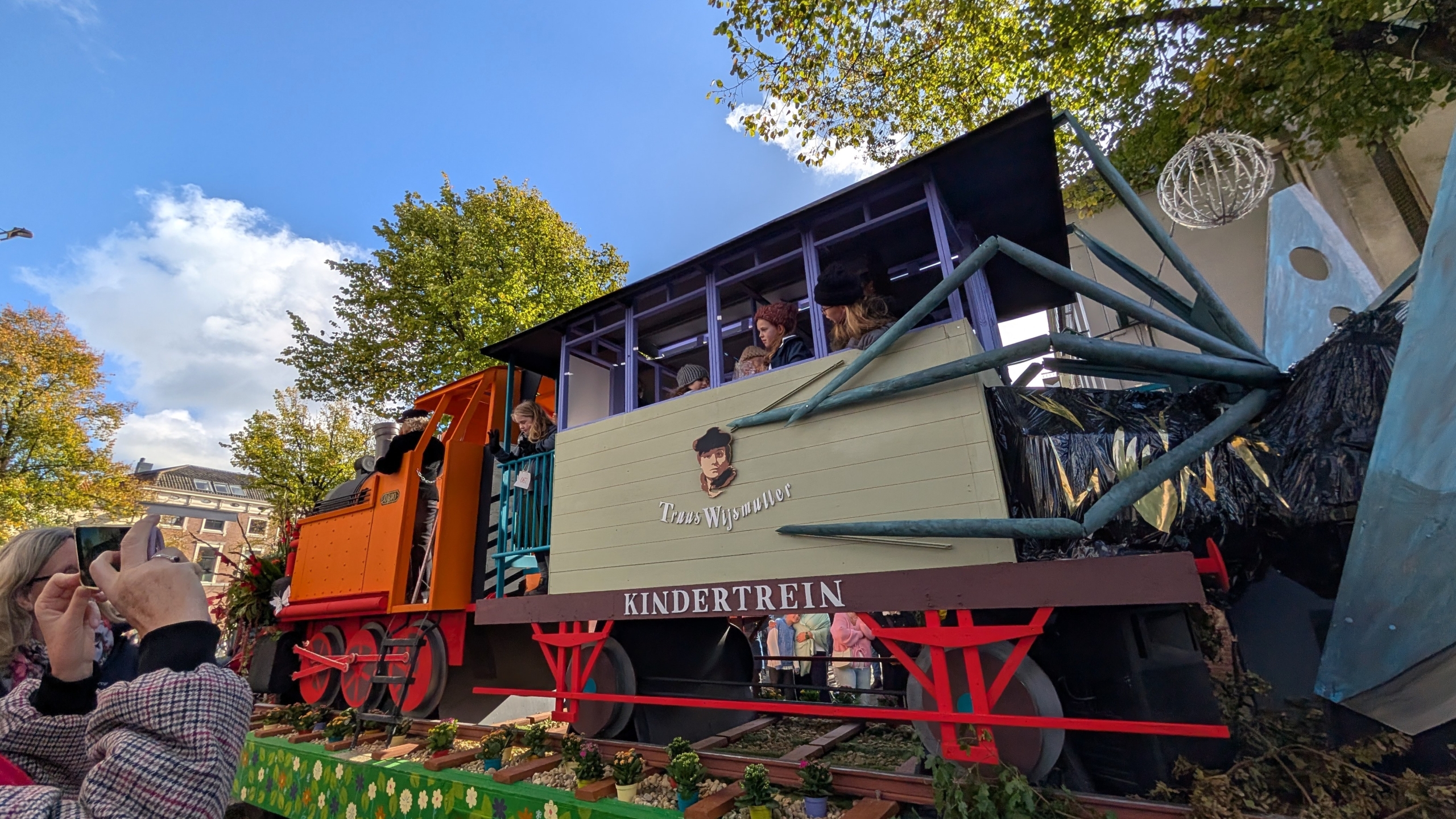 Praalwagen met kindertrein en passagiers, met op de zijkant de tekst “KINDERTREIN”, onder een blauwe lucht