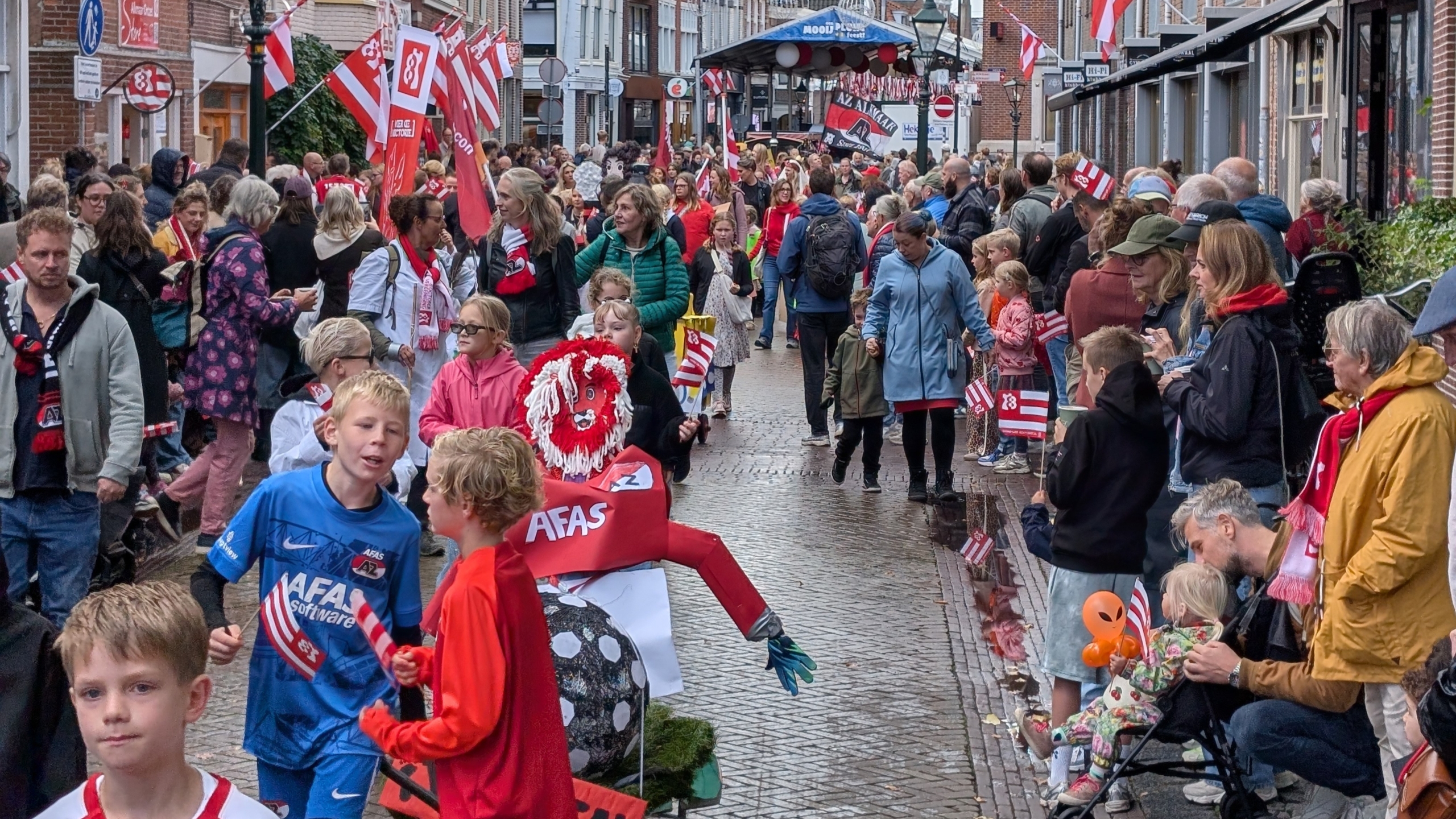 Drukke straat vol supporters met rood-witte AZ-vlaggen en sjaals; kinderen lopen langs een praalwagen met voetbalfiguur