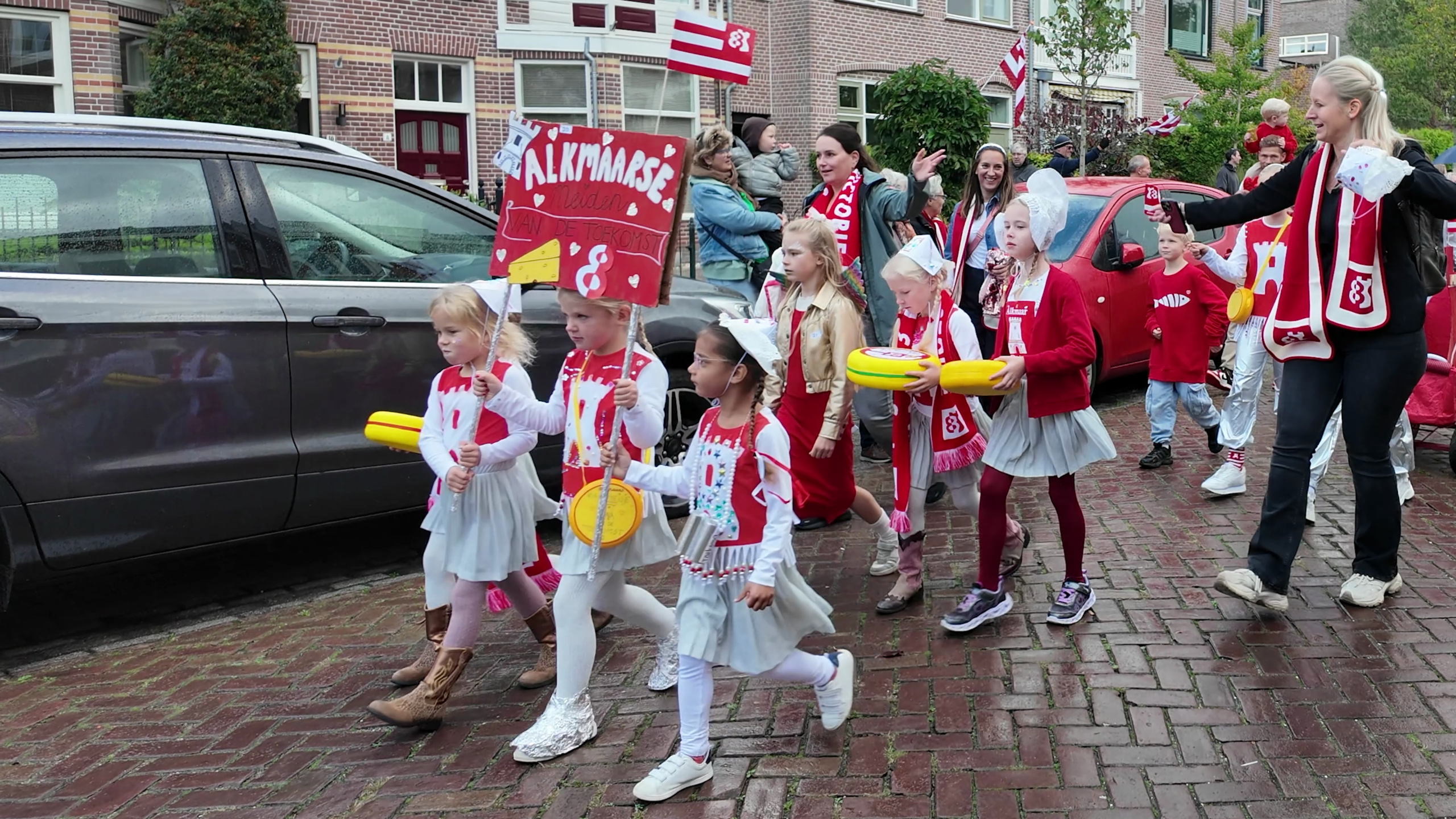 Kinderen in rood-witte outfits lopen in optocht door de straat met vlaggen, kazen en een bord met “Alkmaarse”