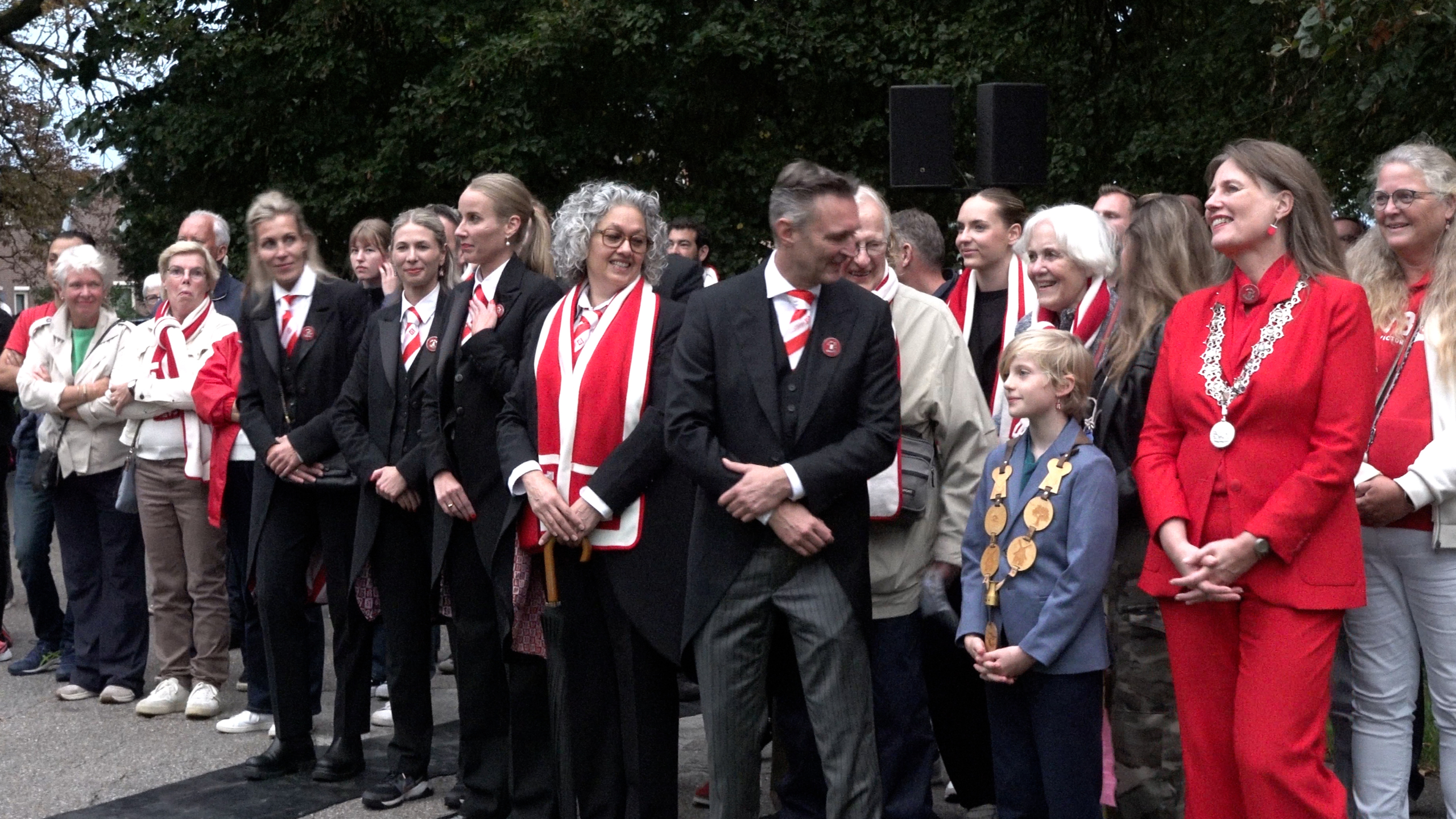 Groep mensen bij een buitenevenement, sommigen in zwarte pakken met rood-witte sjaals en een vrouw in rood met ambtsketen