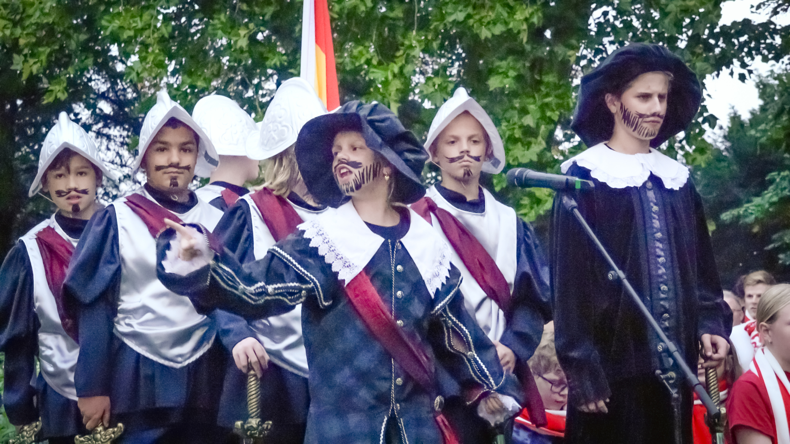 Groep kinderen in historische musketierskostuums met nepbaarden, sommige met helmen, op een podium in het groen