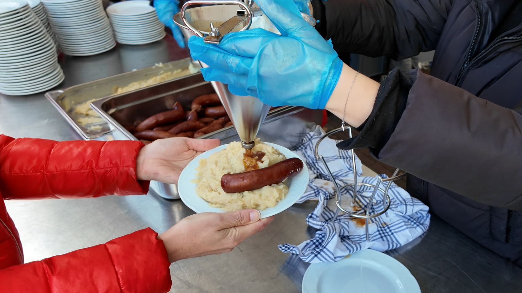 Iemand met blauwe handschoenen spuit saus op zuurkool met worst op een bord bij een uitgiftebalie