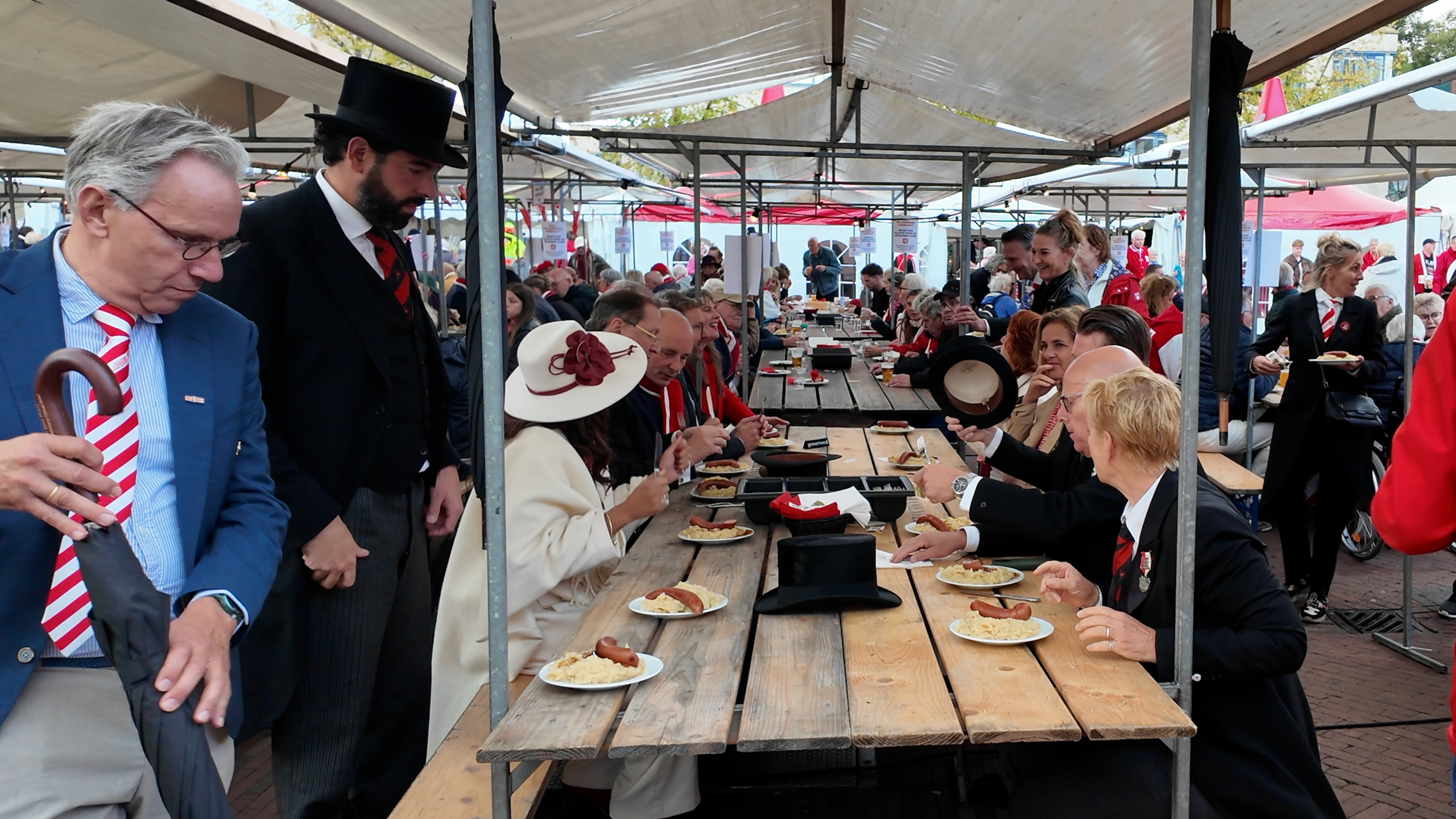 Drukke festivaltent met mensen aan lange houten tafels, borden met worst en eten, sommigen in formele kleding en hoge hoed