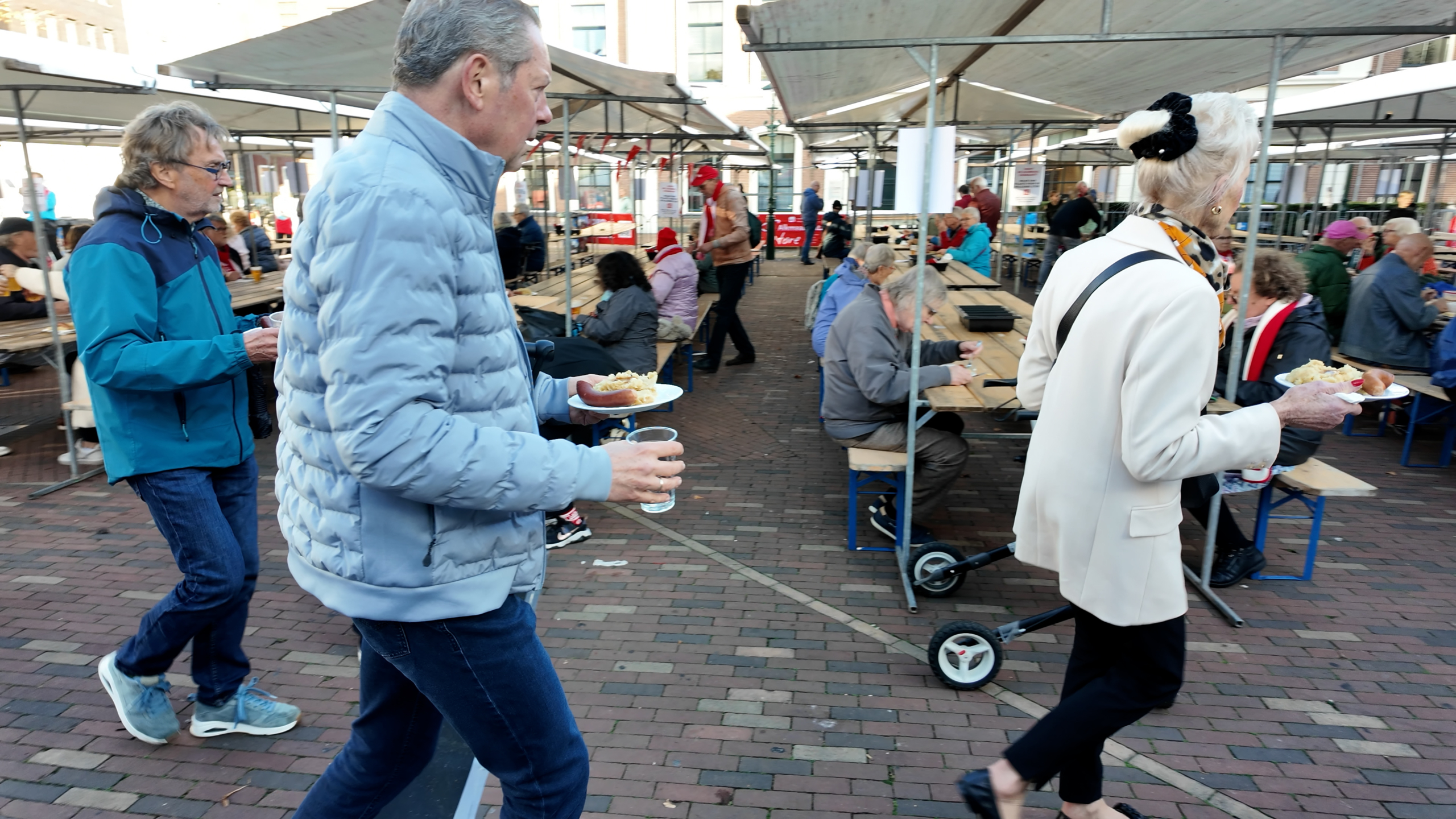 Mensen lopen met borden eten en drankjes langs lange tafels onder tenten op een drukke markt