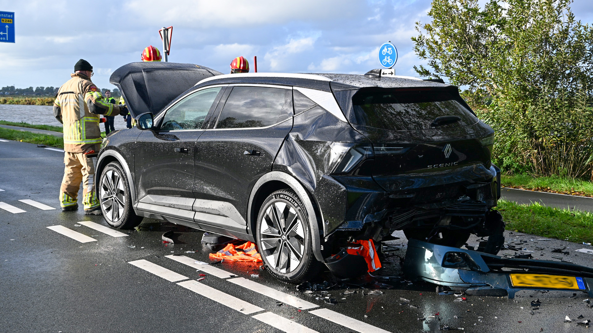 Beschadigde zwarte auto na aanrijding op natte weg, met brandweer en brokstukken achter de auto