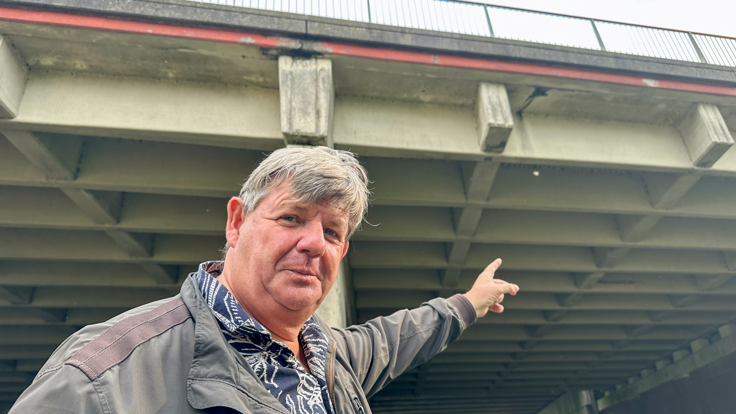 Man wijst omhoog naar de onderkant van een betonnen brug, met zichtbare balken en beschadigingen aan de rand