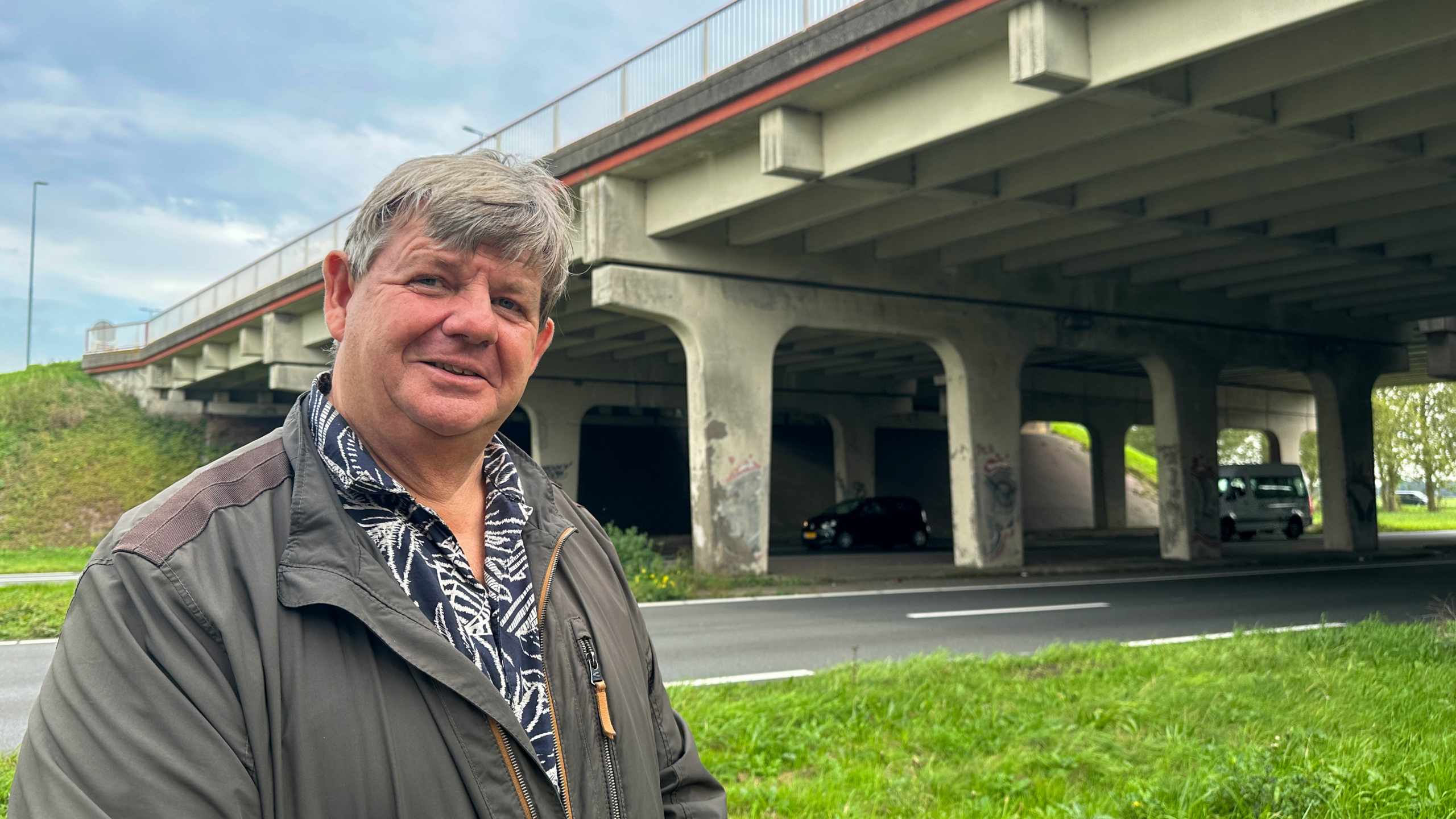 Man met grijs haar staat bij een viaduct; onder de brug zijn betonpijlers, graffiti en passerende auto’s te zien