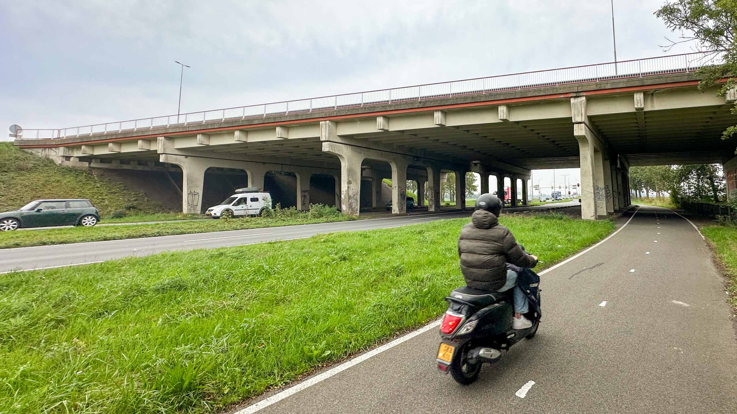 Scooterrijder op fietspad naast een weg onder een betonnen viaduct, met auto’s en graffiti op de pijlers