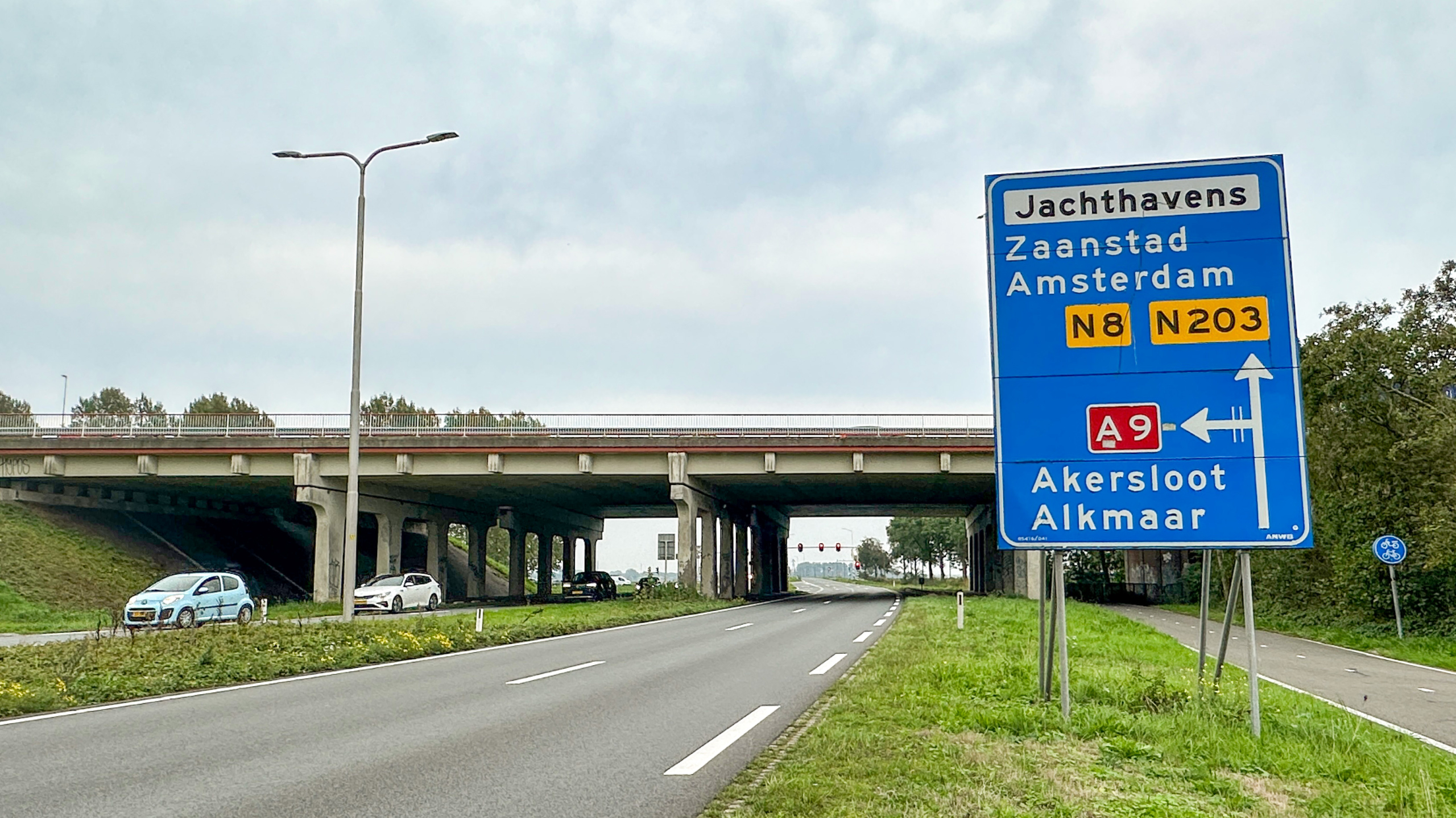 Blauw verkeersbord bij viaduct met verwijzingen naar Zaandstad, Amsterdam (N8/N203) en A9 naar Akersloot en Alkmaar
