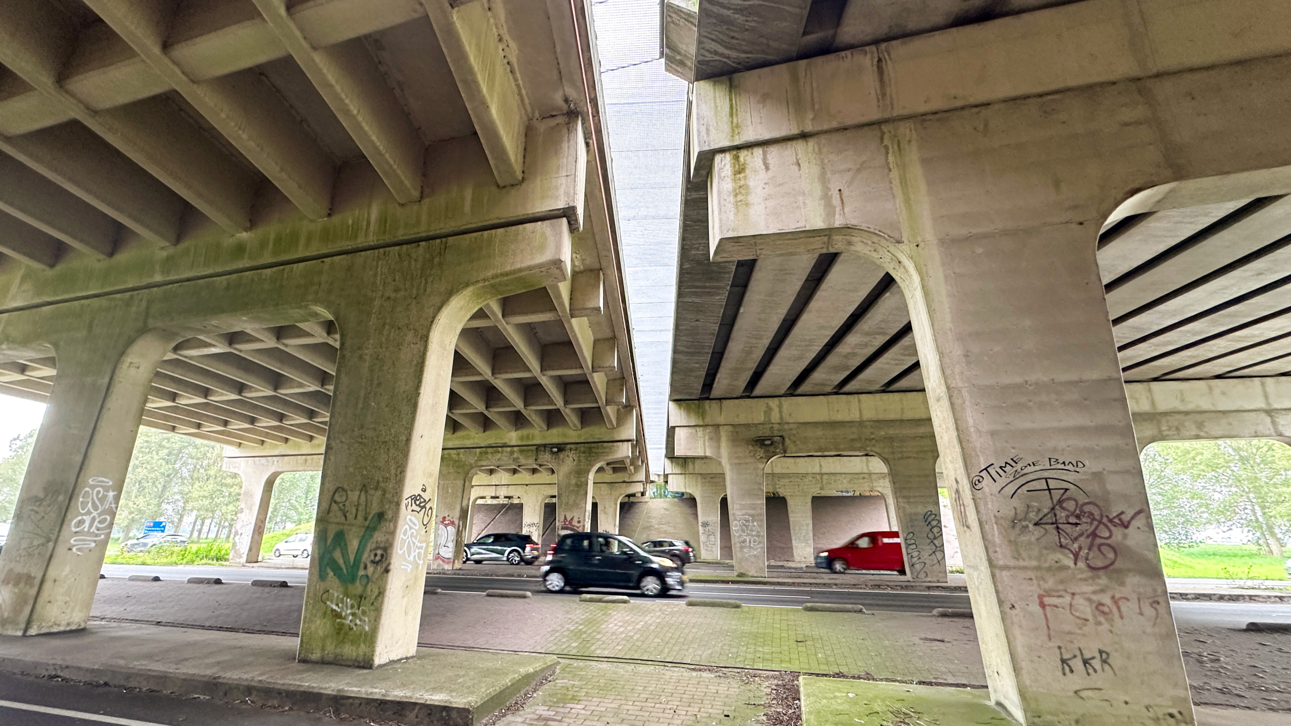 Onderzijde van een betonnen viaduct met pilaren vol graffiti en auto’s die eronder rijden