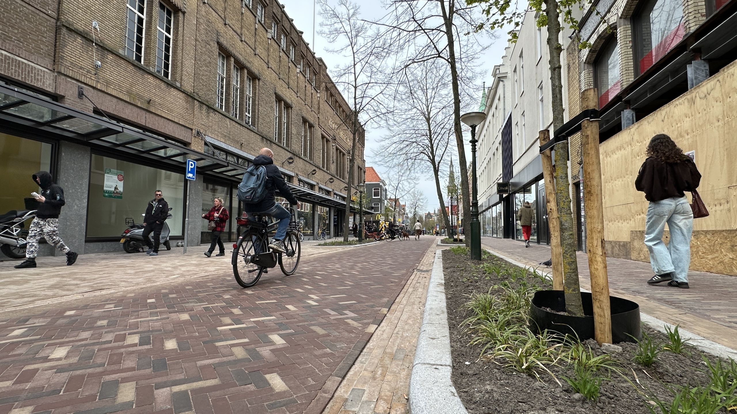 Stadsstraat met fietser en voetgangers langs winkels, jonge bomen en bloemperk, met bouwschutting rechts