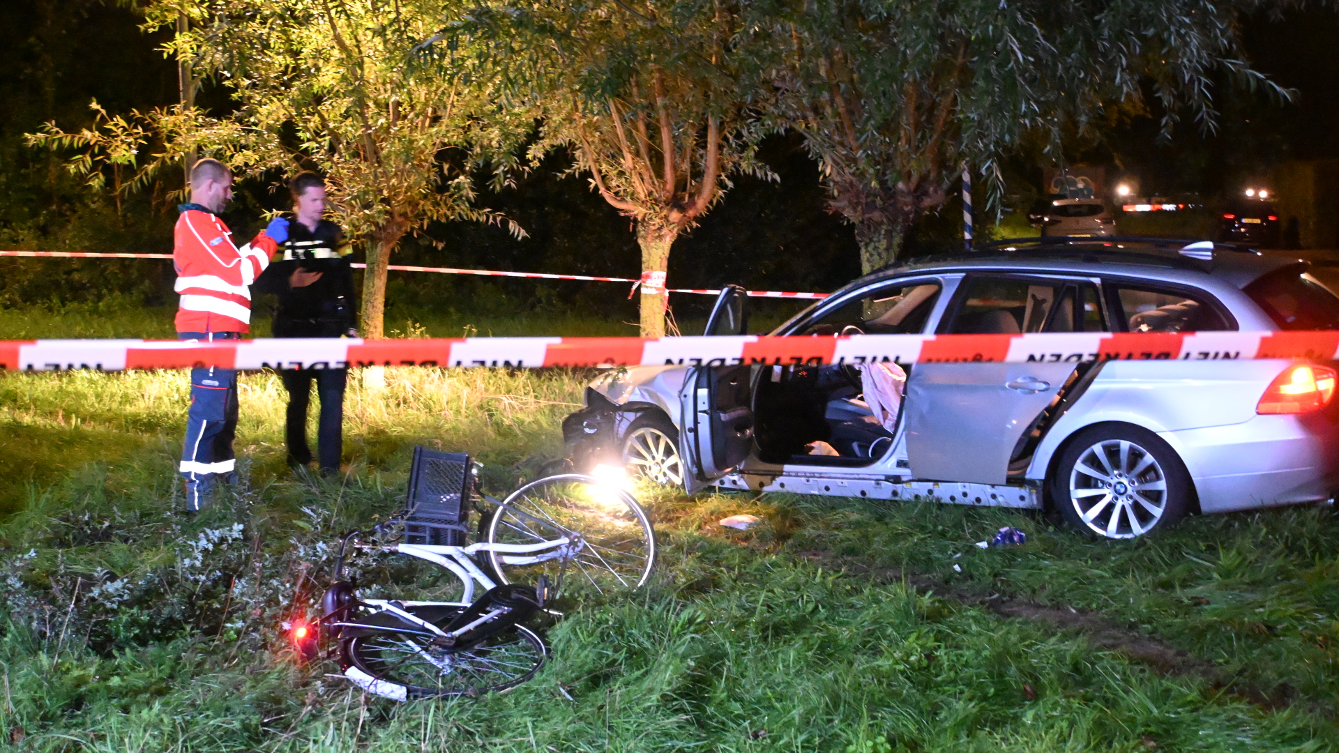 Afgezette ongevalslocatie met beschadigde auto en omgevallen fiets in het gras, met hulpverleners in de achtergrond