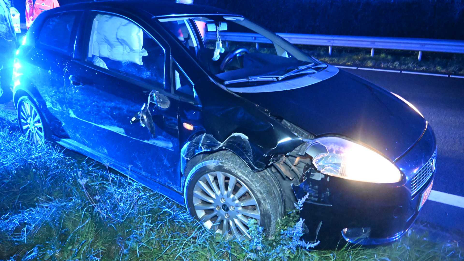 Zwarte auto na aanrijding langs de weg, met uitgeklapte airbag en schade aan de linker voorkant, in blauw zwaailicht