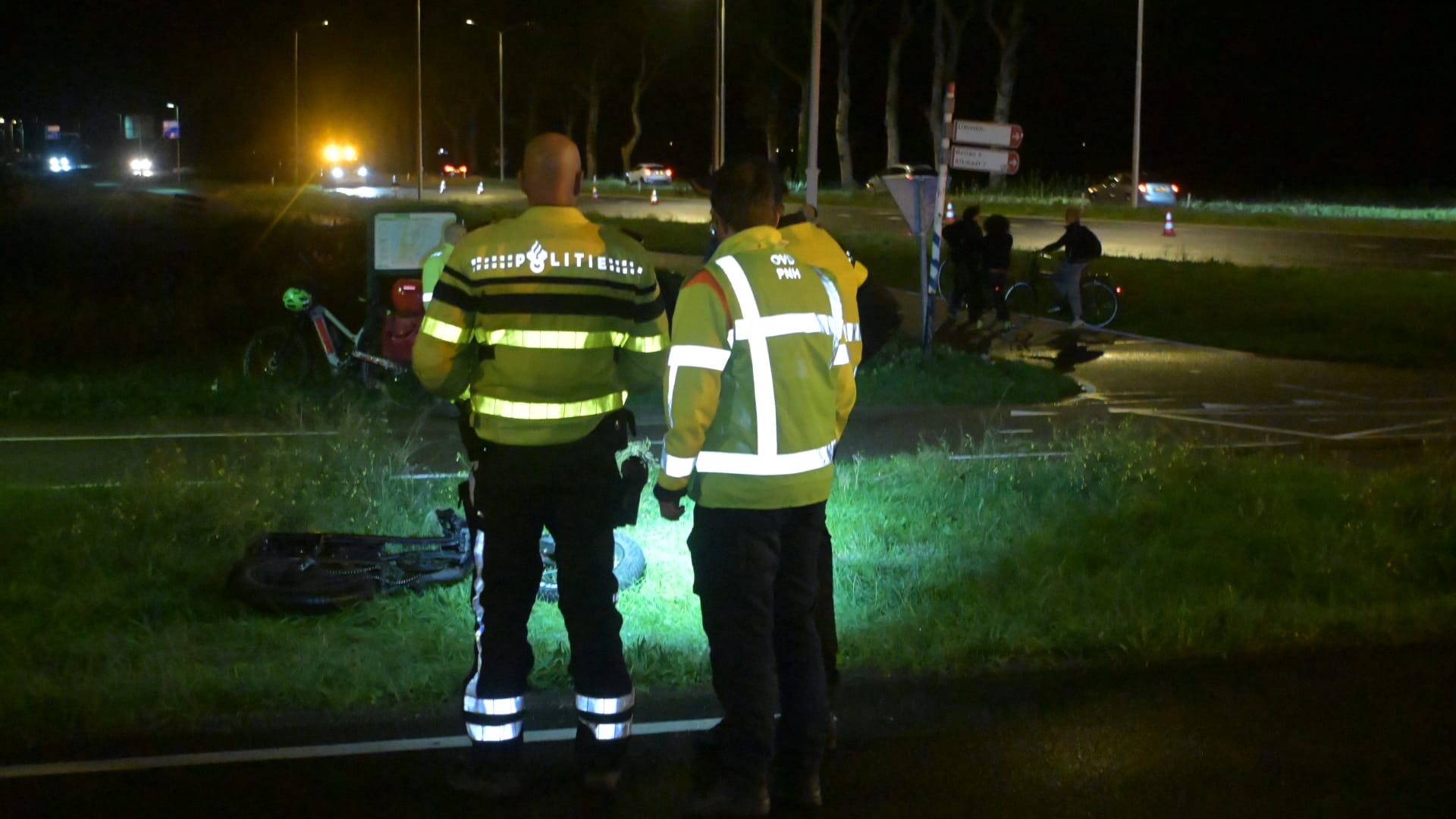Politie en hulpverleners onderzoeken ’s nachts een ongevalslocatie langs de weg, met een omgevallen fiets in het gras