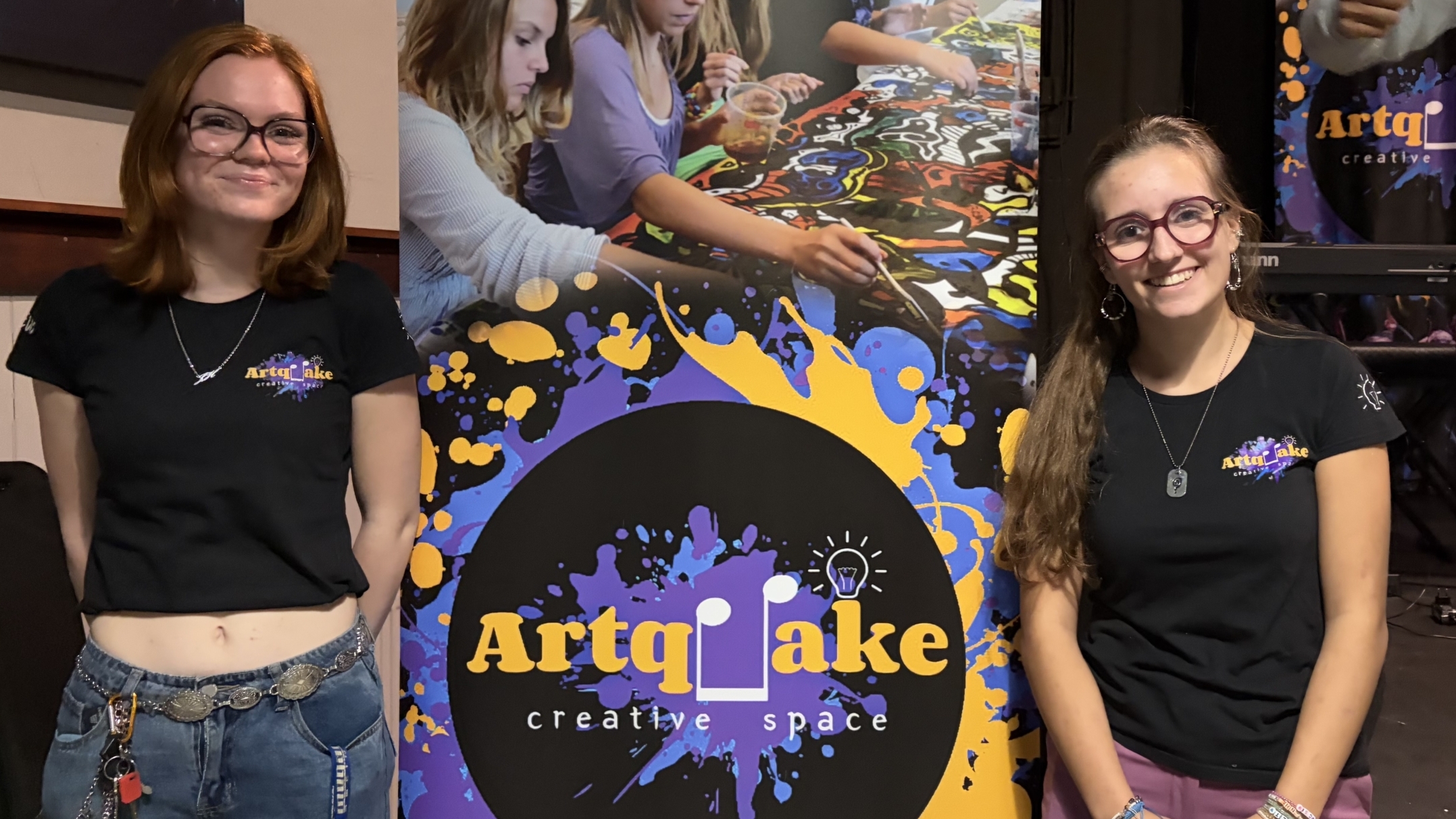 Twee vrouwen in ArtqLake‑shirts poseren glimlachend bij een kleurrijke ArtqLake creative space‑banner