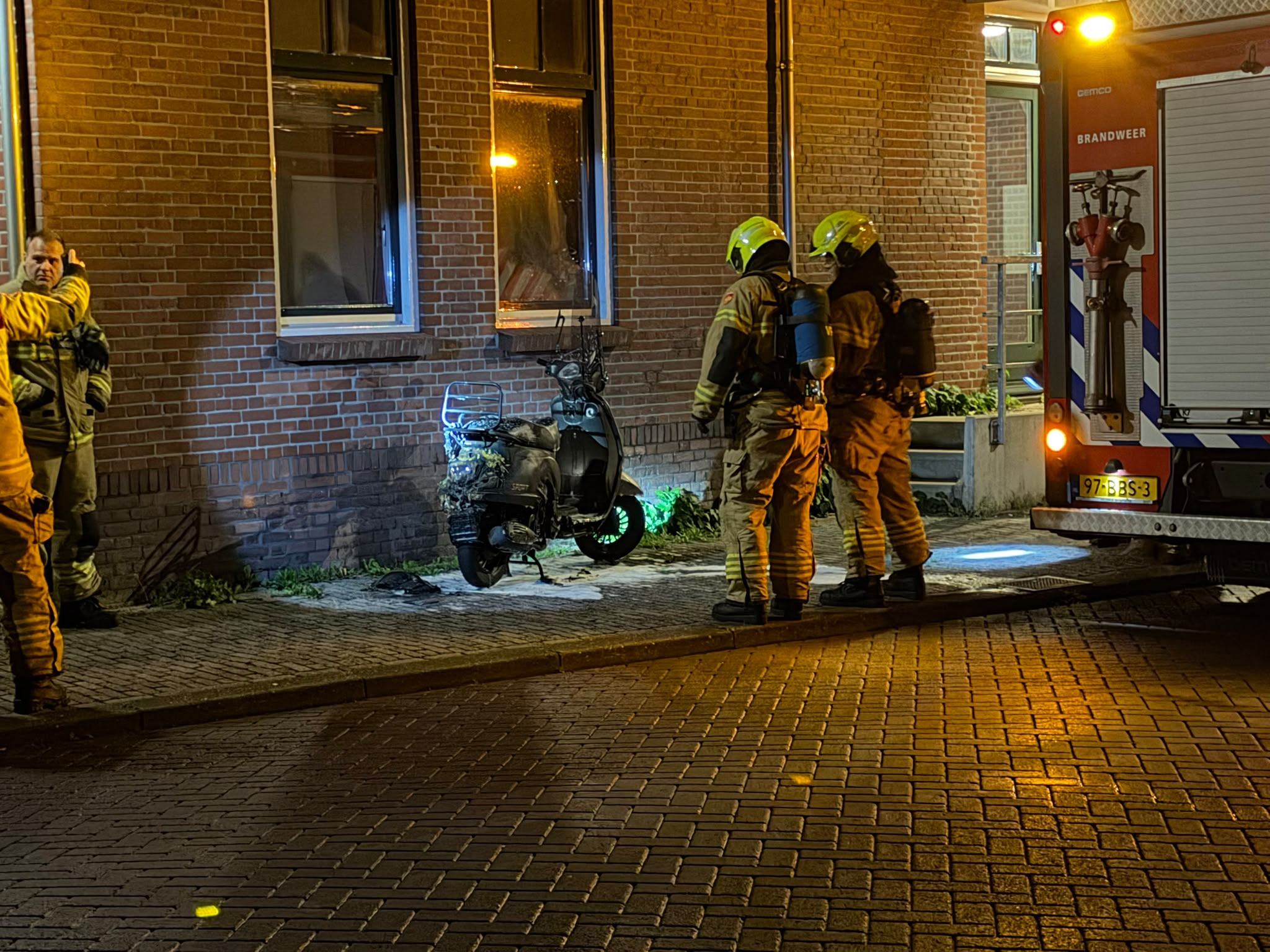 Brandweerlieden bij een bakstenen gebouw naast een brandweerwagen, inspecteren een uitgebrande scooter op straat