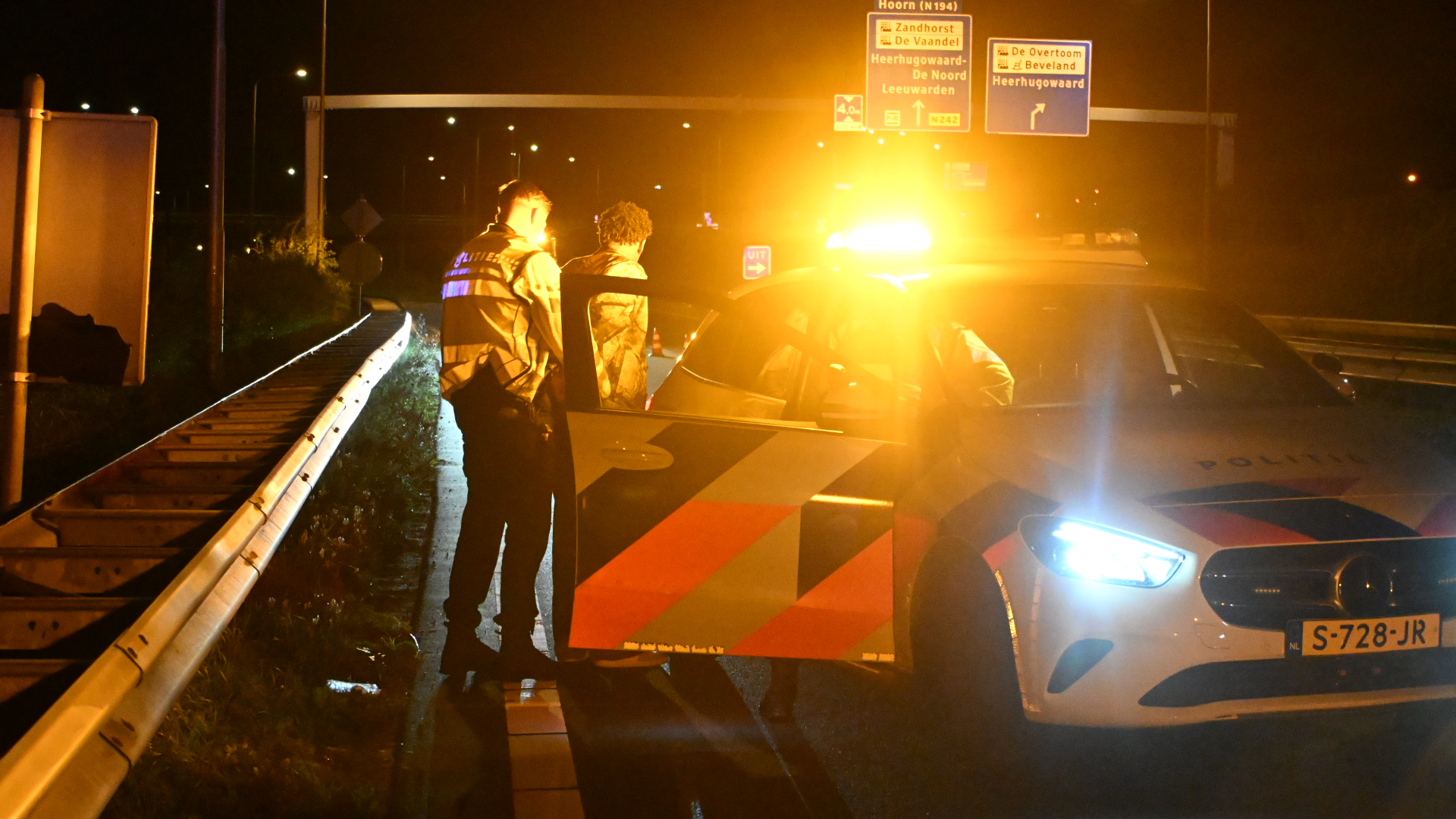 Politieagent bij een man naast een politieauto met knipperlichten langs de snelweg, met richtingborden op de achtergrond