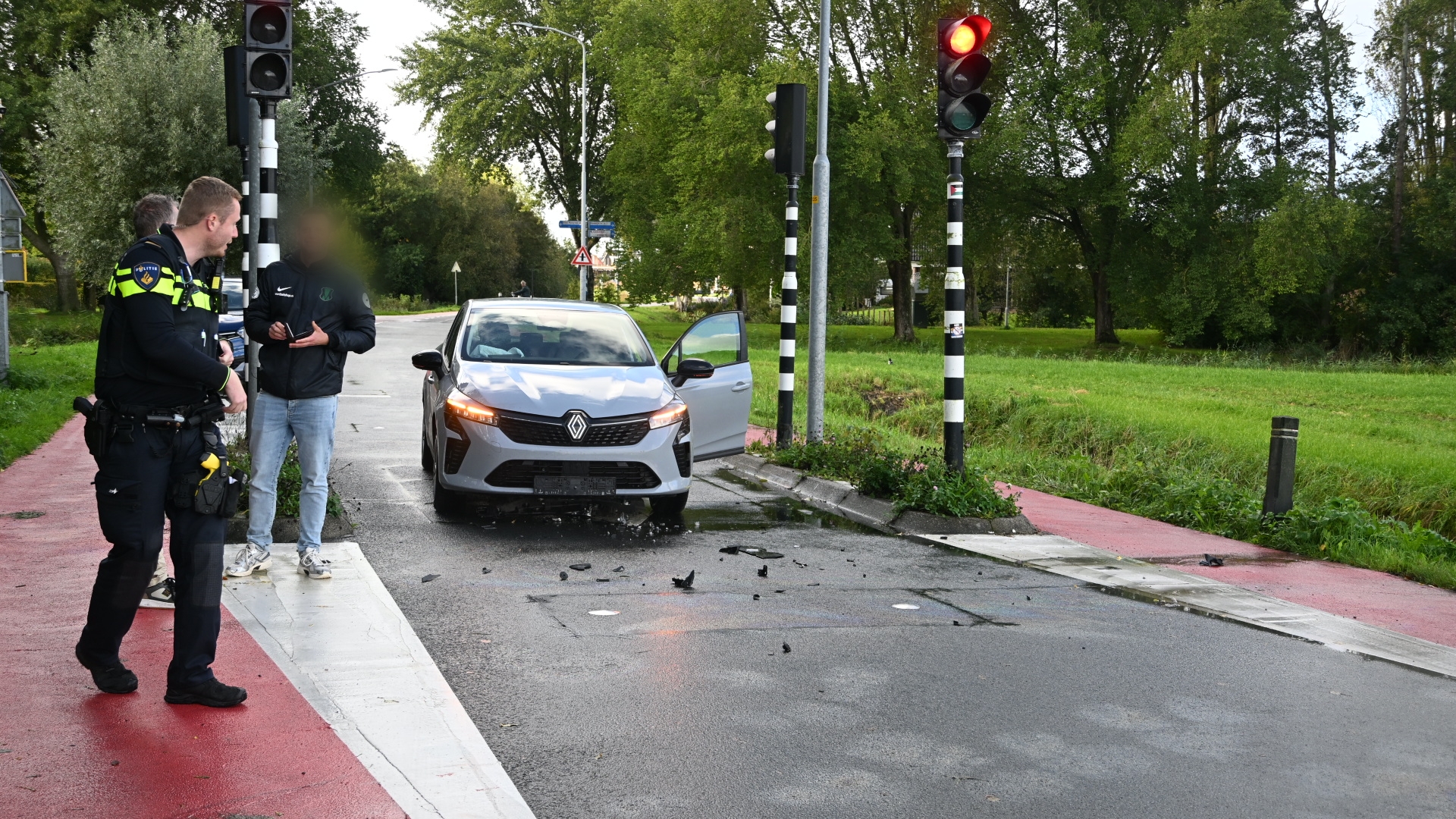 Politieagent praat met bestuurder naast grijze auto met open portier bij kruispunt met rood licht en brokstukken op weg
