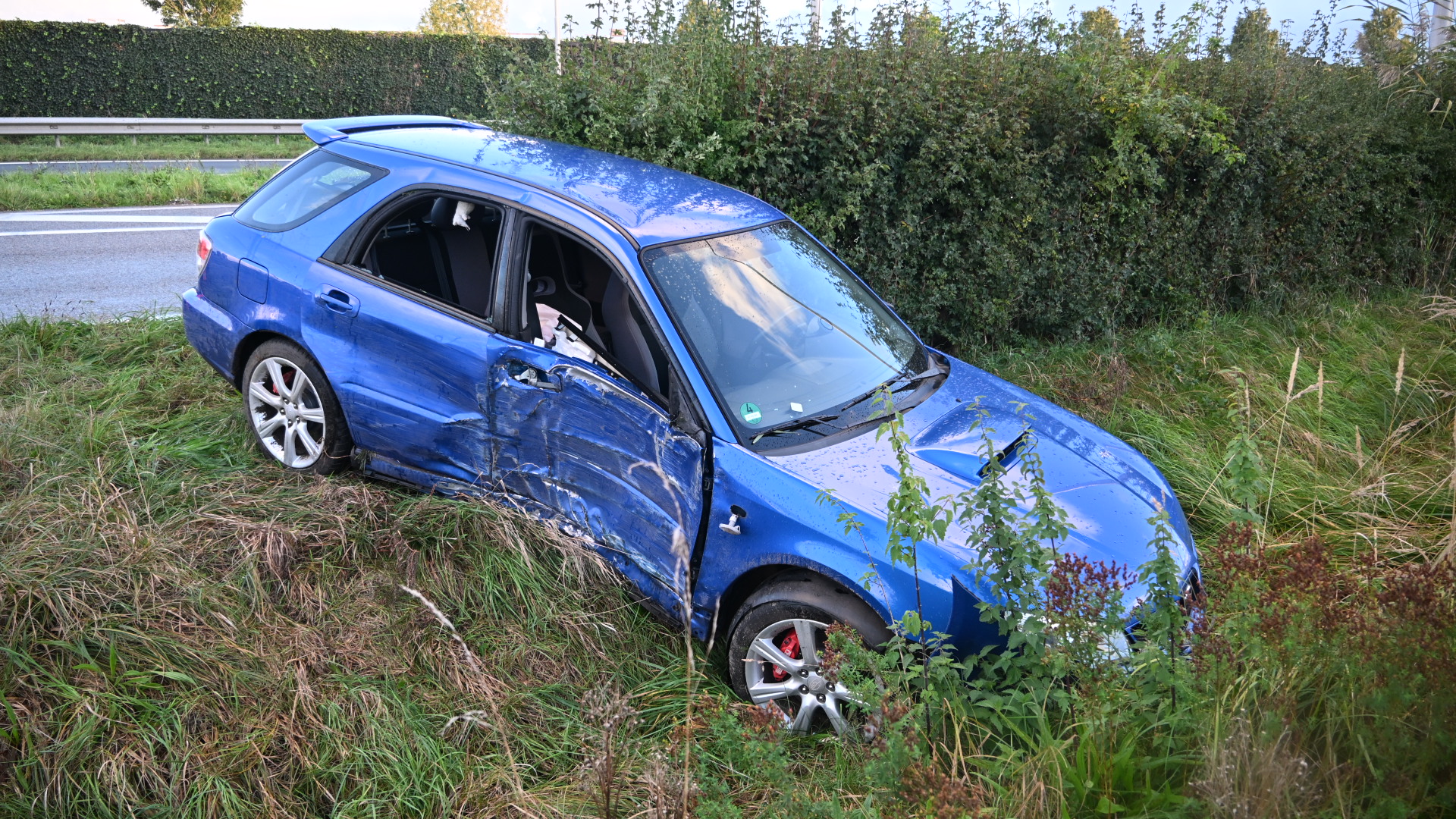 Blauwe auto met schade aan de zijkant staat deels in de berm naast een weg, omringd door gras en struiken