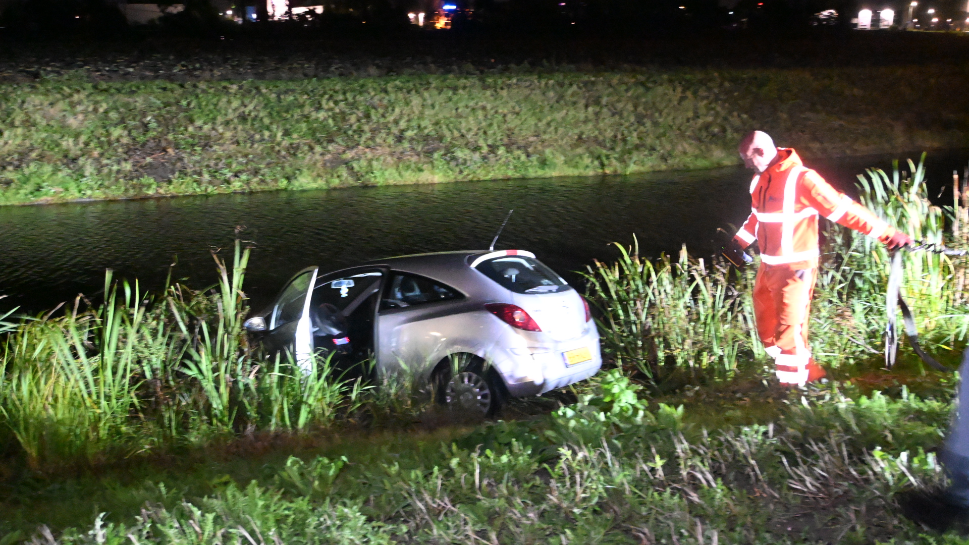 Auto in de berm langs een sloot, met een hulpverlener in oranje reflectiekleding bij het voertuig in de nacht