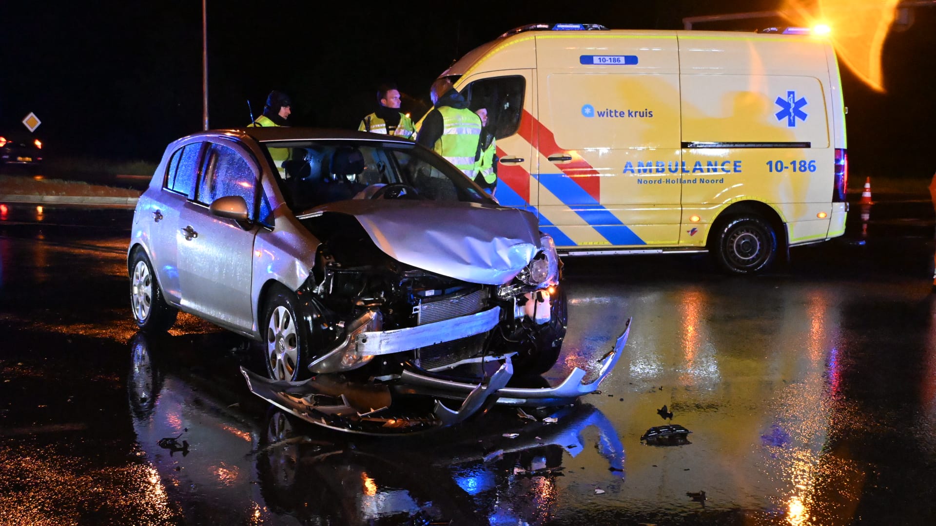 Zwaar beschadigde auto op nat wegdek na aanrijding, met ambulance en hulpverleners op de achtergrond