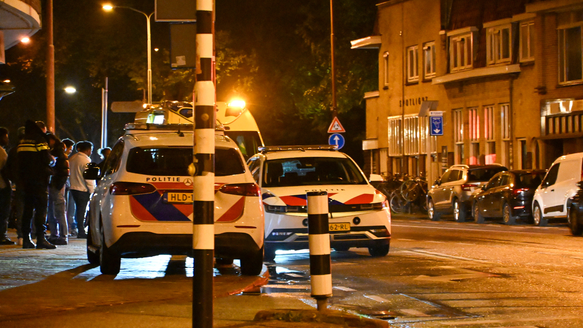 Twee politieauto’s met zwaailichten op een natte straat in de avond, met omstanders op de stoep