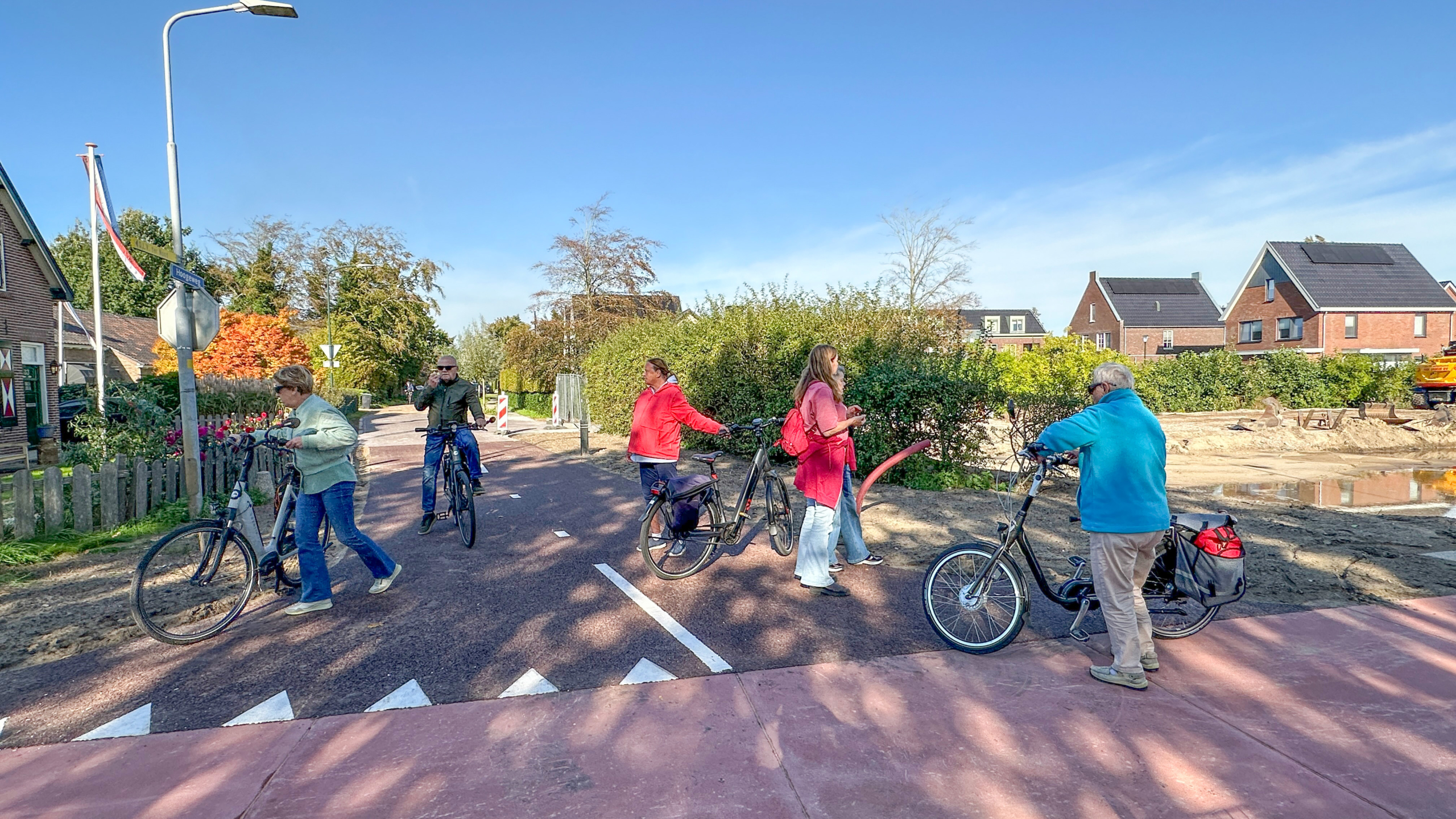 Vier fietsers staan stil op een fietspad bij wegwerkzaamheden, met huizen en bomen op de achtergrond onder een blauwe lucht