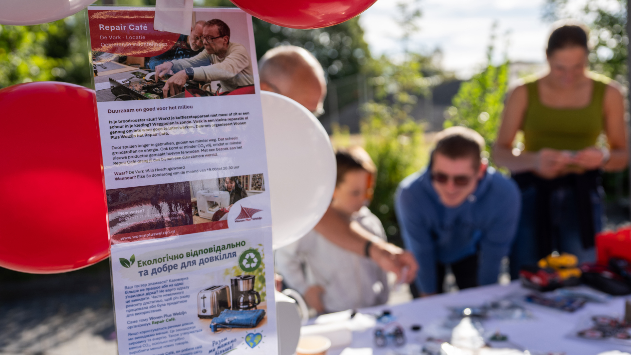 Flyer voor Repair Café met rode en witte ballonnen, met op de achtergrond onscherpe bezoekers aan een tafel buiten