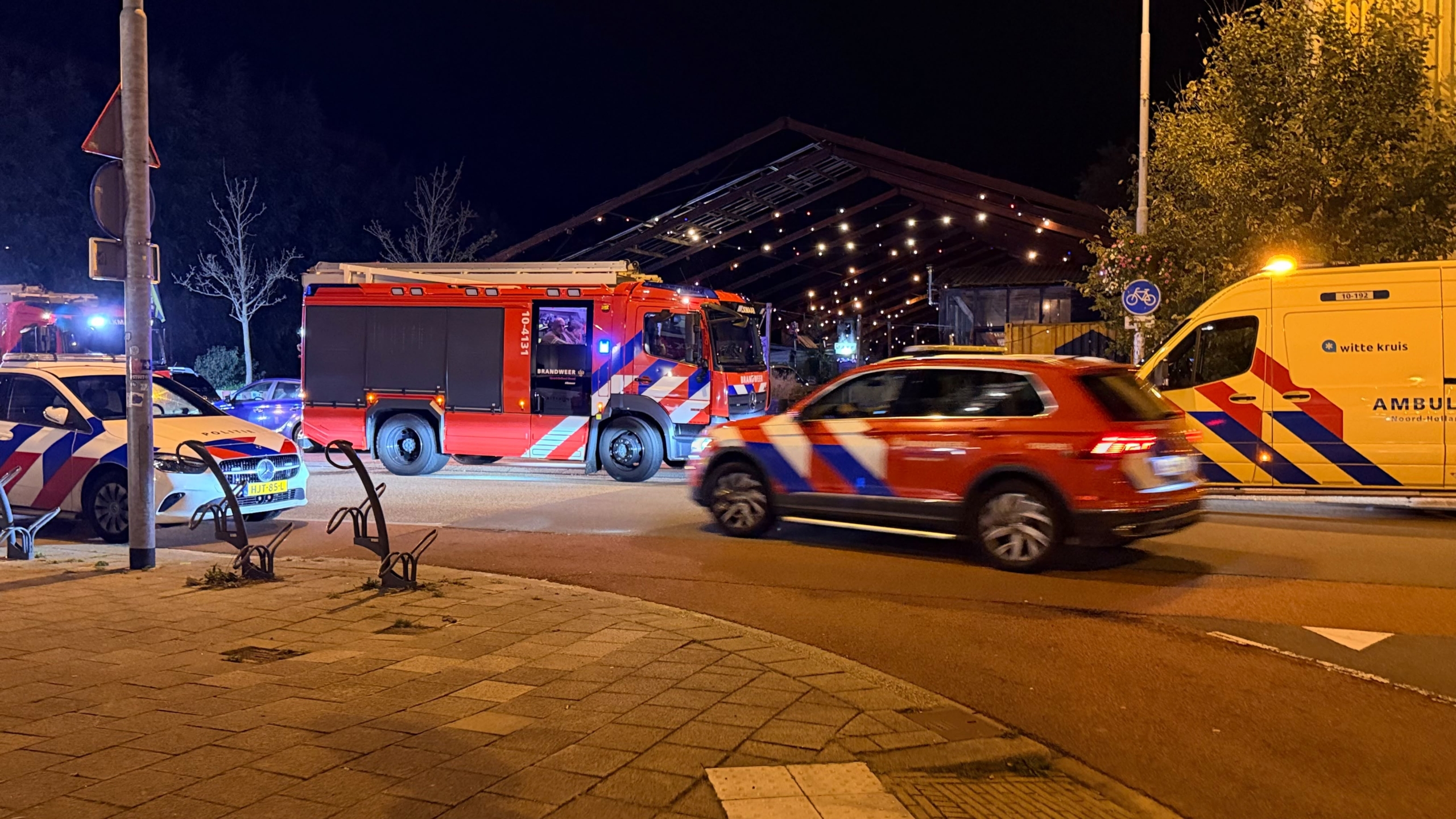 Brandweerwagen, politieauto en Witte Kruis-ambulance met zwaailichten bij een verlicht gebouw in de nacht
