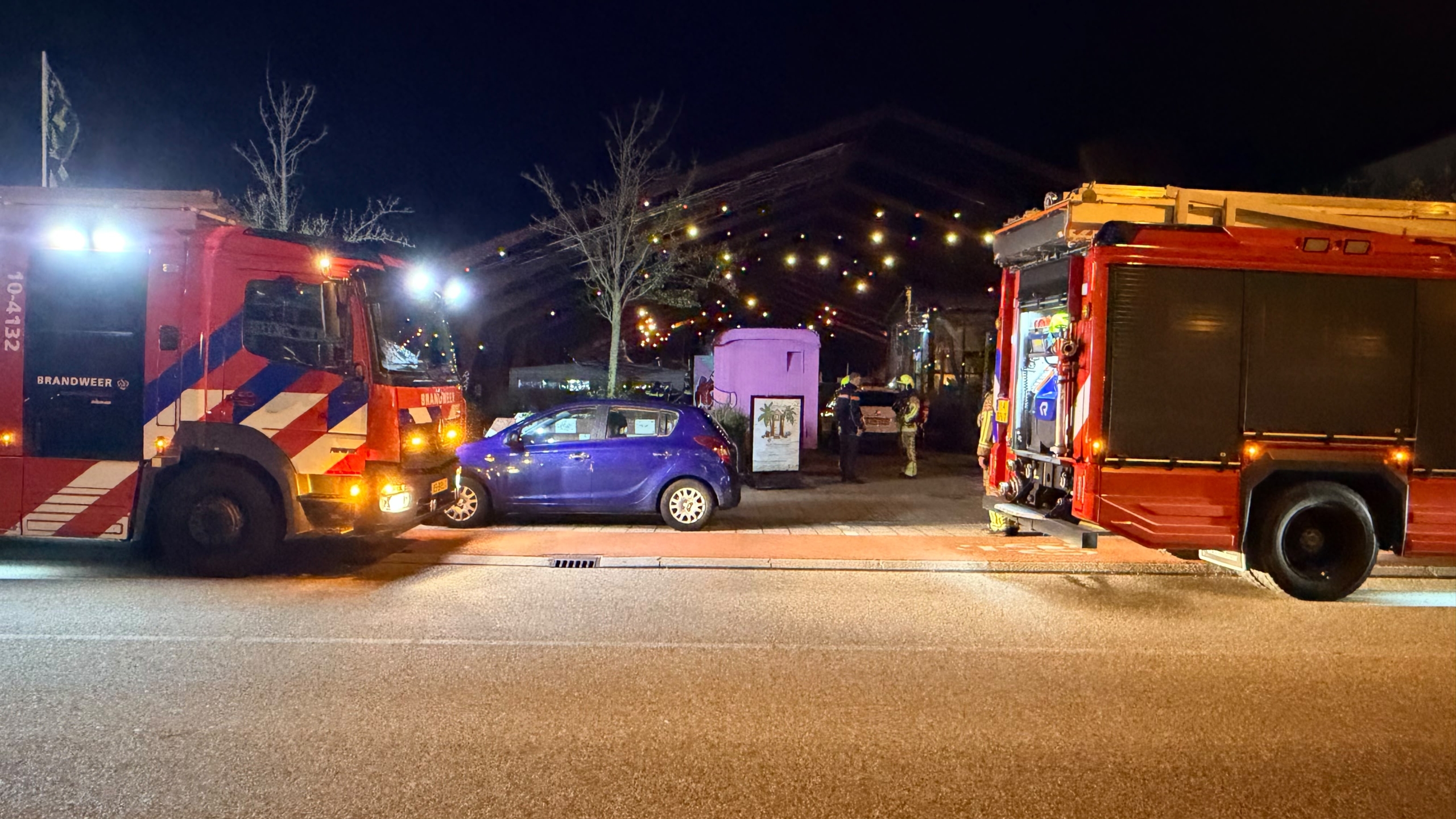 Twee brandweerwagens met zwaailichten bij een gebouw in de nacht, met een paarse auto en brandweerlieden ertussen