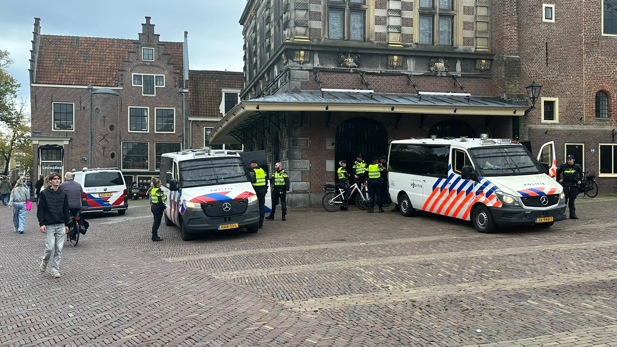 Politieagenten bij drie politiebusjes voor een historisch gebouw op een stadsplein met voorbijgangers
