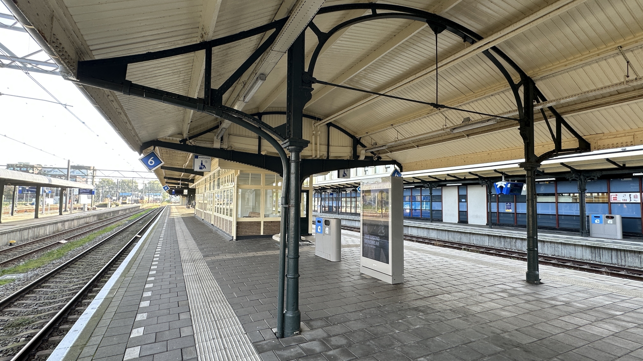Leeg perron 6 op een treinstation met overkapping, spoorrails, prullenbakken en een digitaal informatiebord