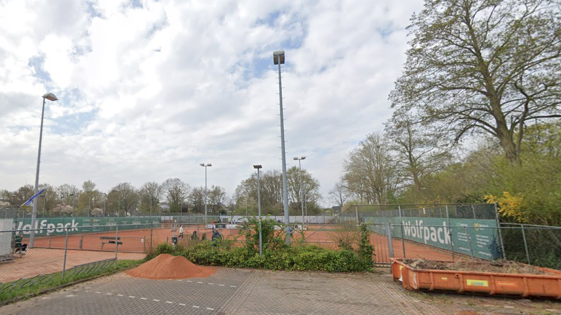 Tennisbanen met gravel, omheind door hekwerk met reclameborden, lantaarnpalen en bomen onder een bewolkte lucht