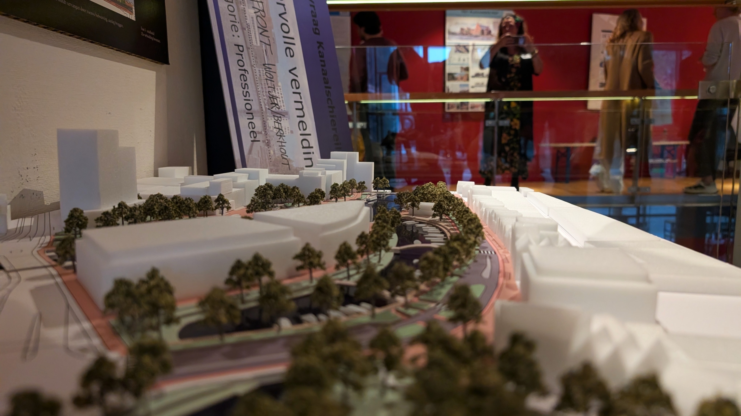 Maquette van een stadsgebied met witte gebouwen, bomen langs een weg en bezoekers op de achtergrond in een tentoonstelling