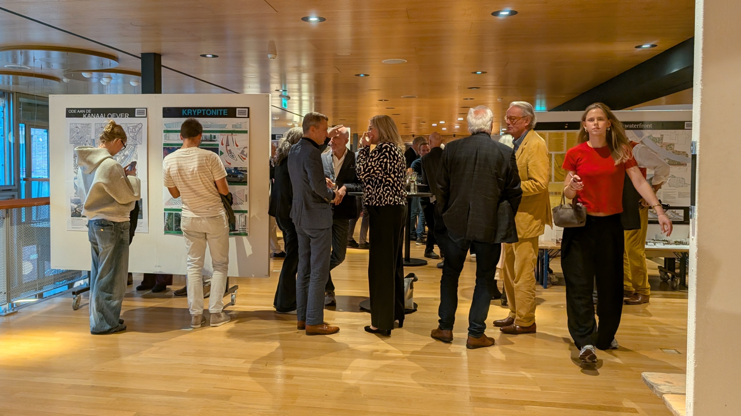 Mensen netwerken op een evenement met posters met kaarten en plannen, terwijl een vrouw in rood langsloopt