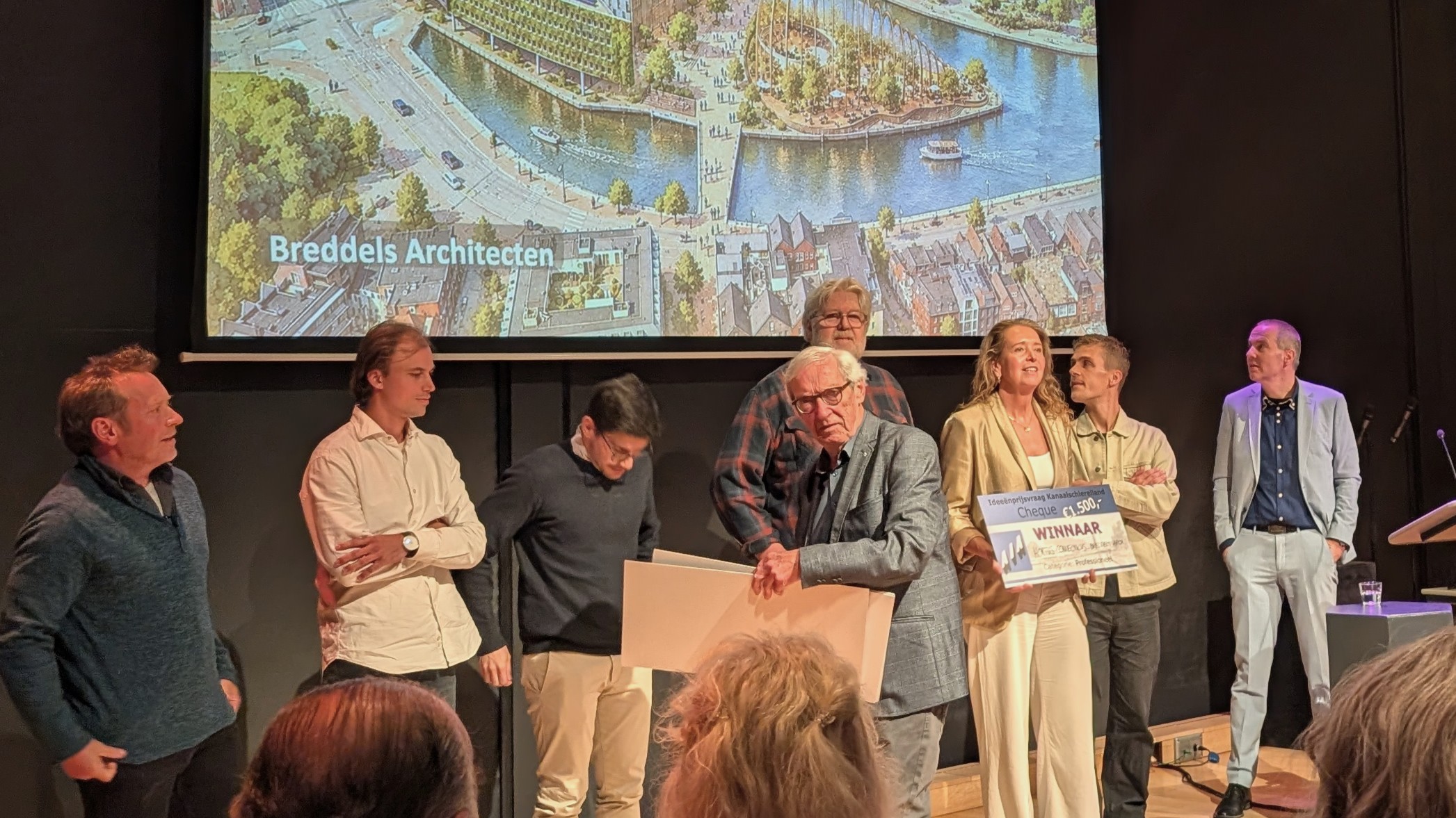 Groep mensen op podium bij prijsuitreiking; winnaar houdt een grote cheque vast, met projectpresentatie op scherm erachter