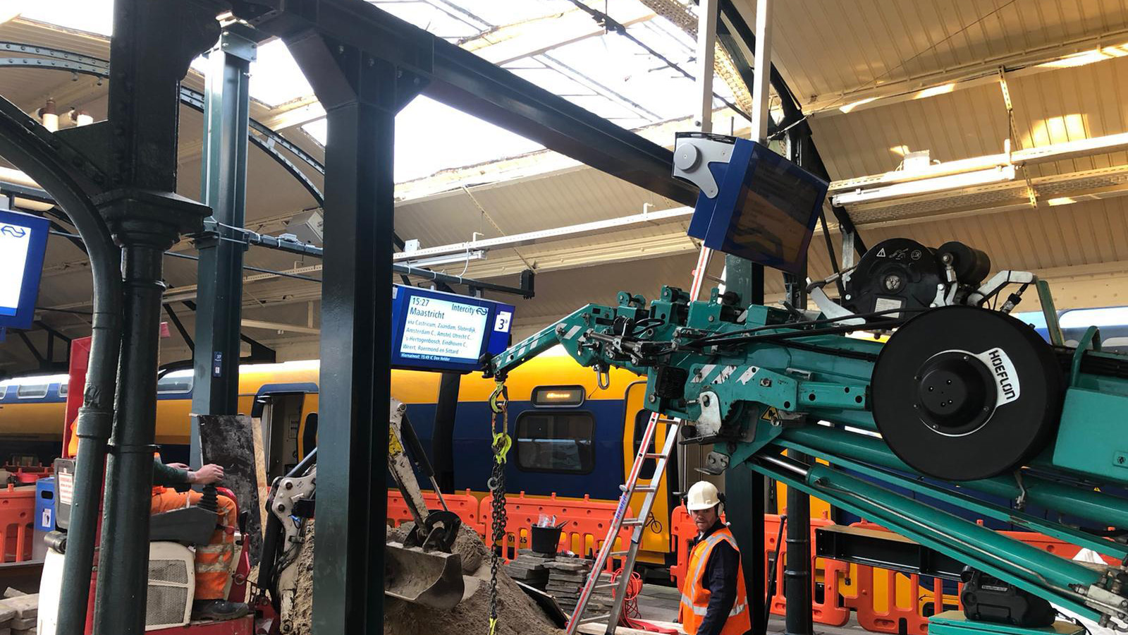 Werkzaamheden op station met bouwmachines en een NS-trein op de achtergrond onder het perronoverkapping