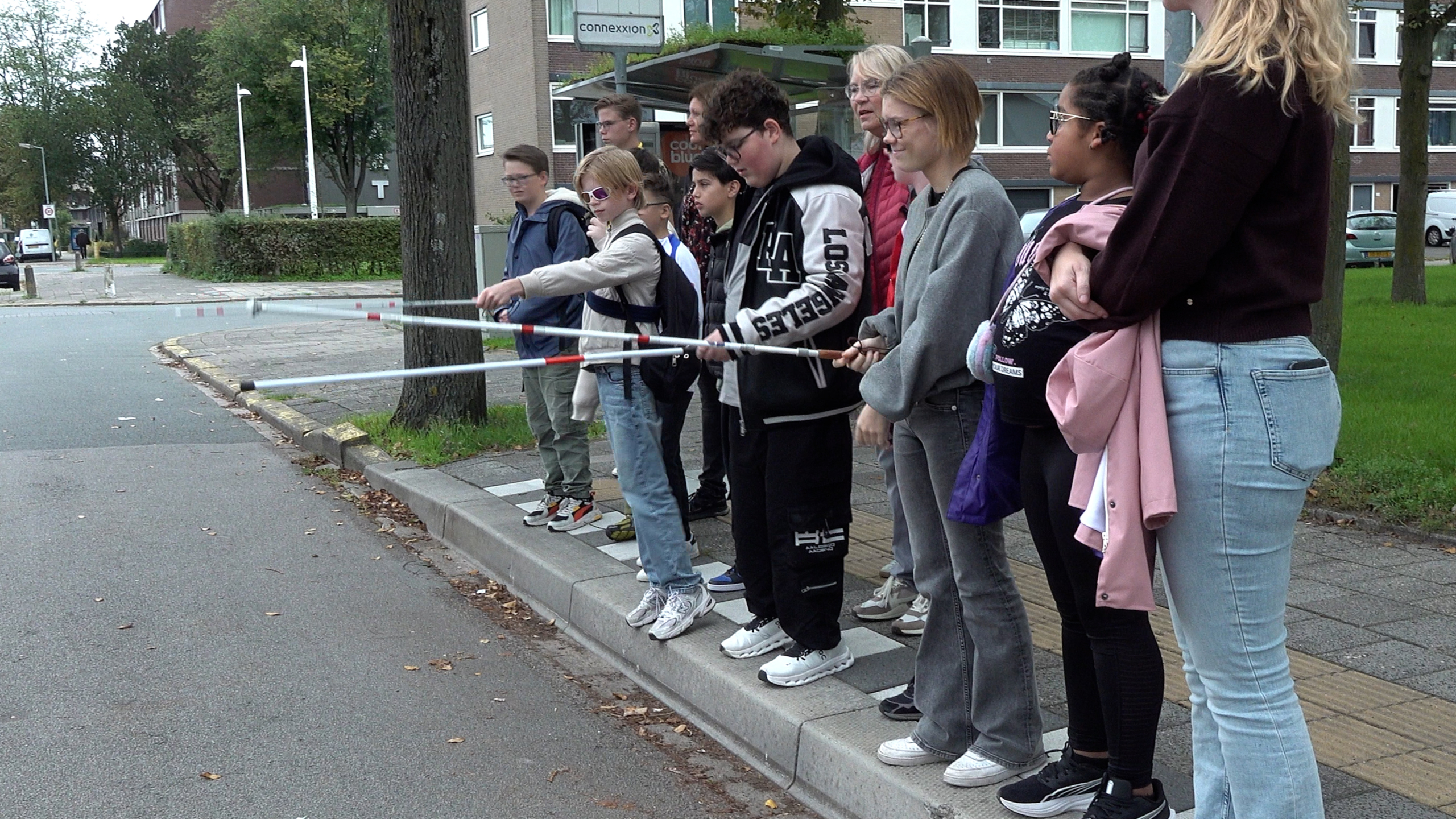 Groep jongeren met blindestokken bij een stoeprand naast een bushalte, klaar om over te steken