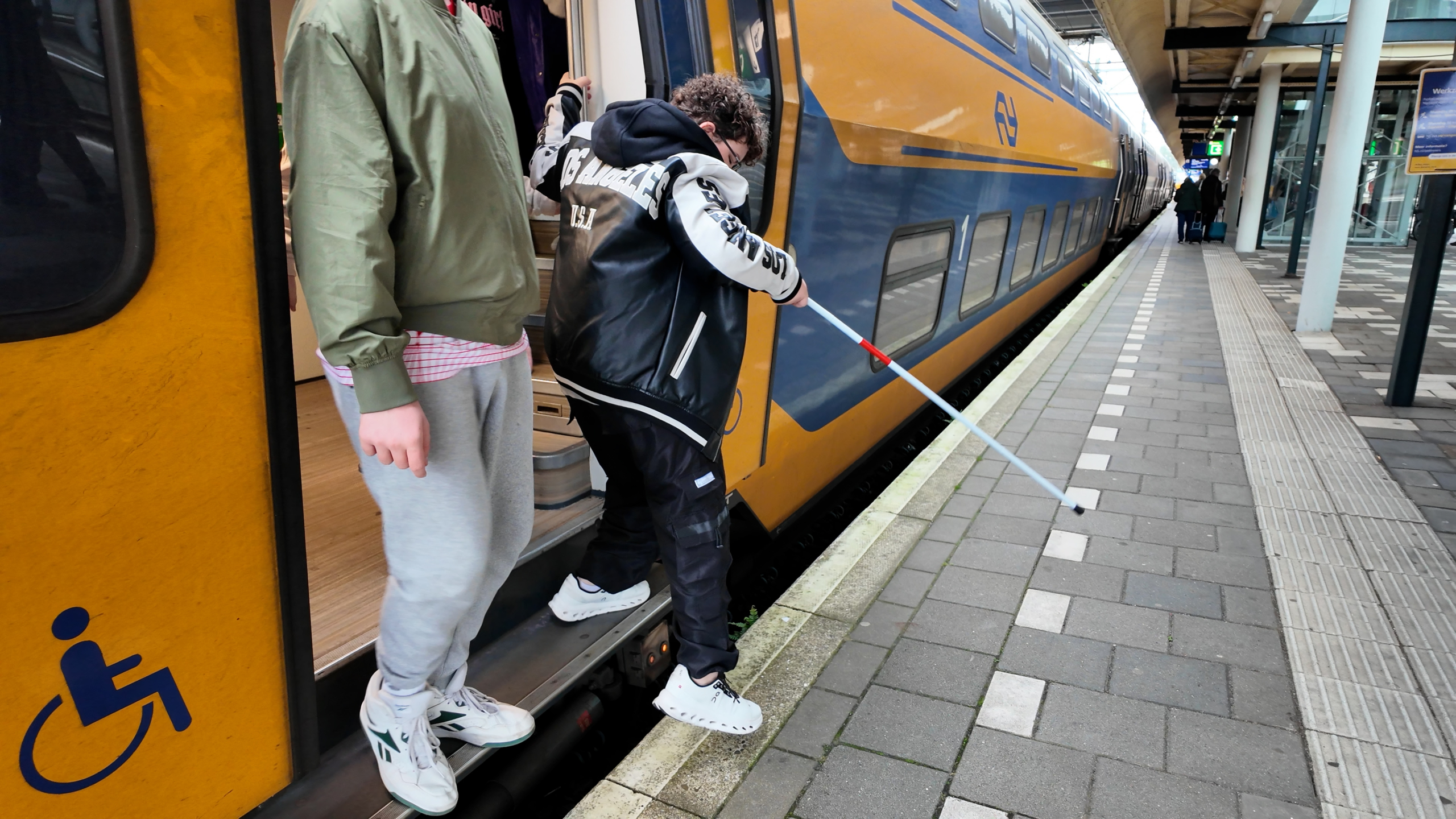 Jongen stapt uit een NS-trein op het perron en houdt een witte stok vast, met rolstoeltoegankelijkheidsicoon op de deur
