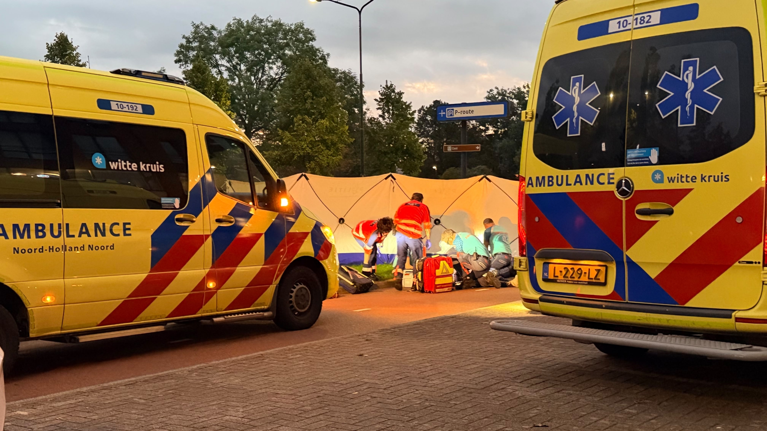 Twee ambulances met hulpverleners die iemand behandelen bij een wit afschermdoek langs de weg in de schemering