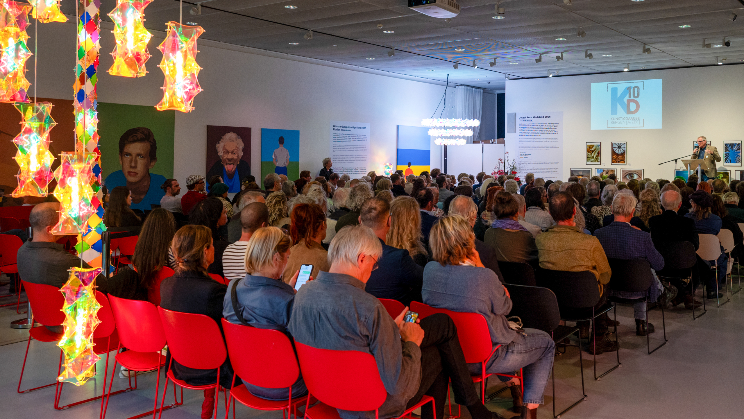 Grote groep bezoekers in een galerie luistert naar een spreker bij podium, met kleurrijke lichtkunst en schilderijen aan de wand