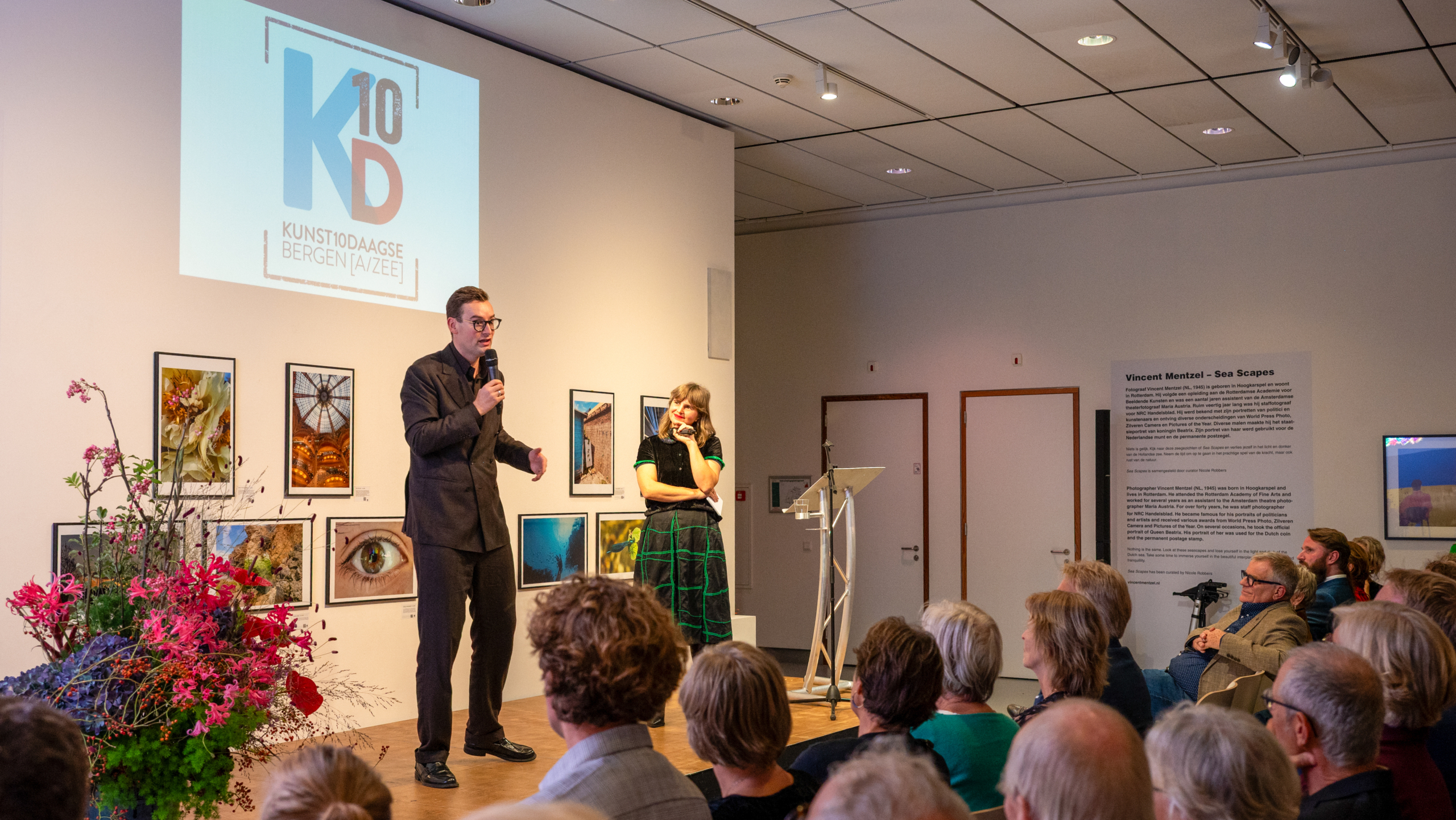 Man spreekt het publiek toe op een podium tijdens Kunst10Daagse Bergen, met projectiescherm en kunstfoto’s aan de muur