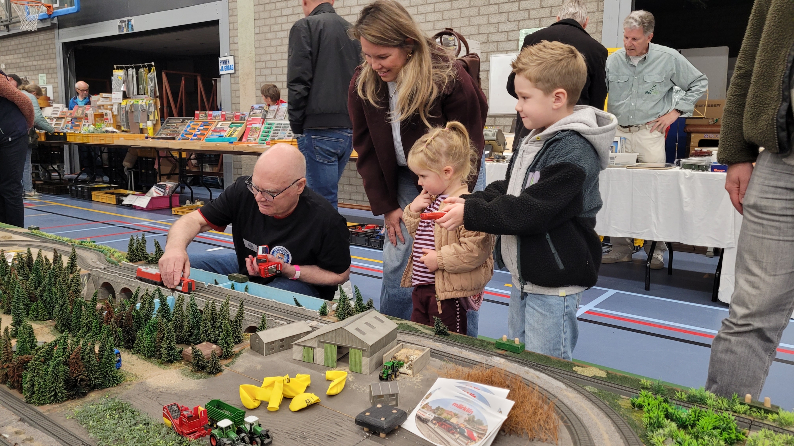 Man laat kinderen een modeltreinbaan bedienen op een beurs, met miniatuurlandschap en tafels met spullen op de achtergrond