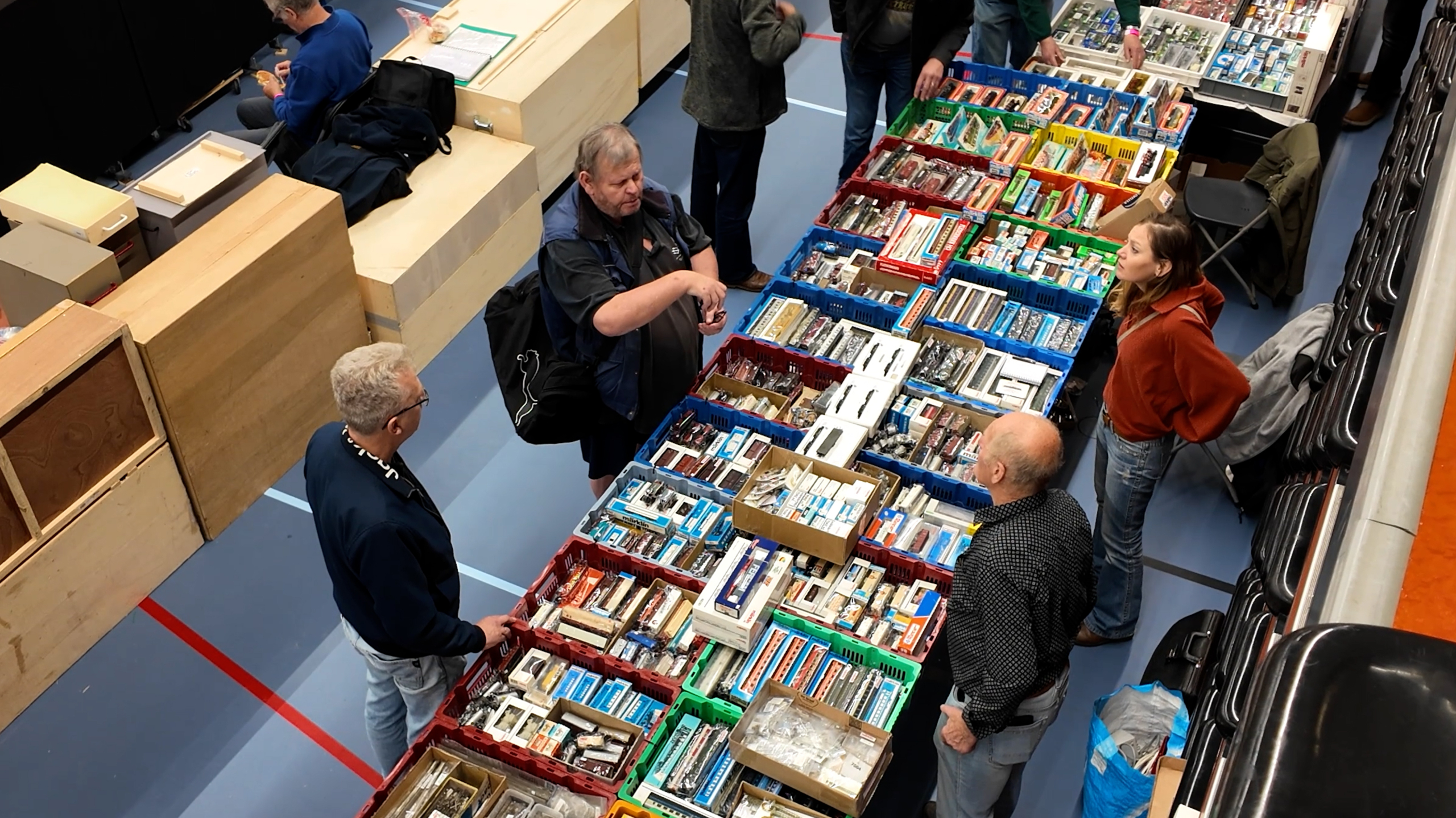 Bezoekers bekijken en bespreken modeltreinen en onderdelen in kratten op een beursvloer, gezien van bovenaf