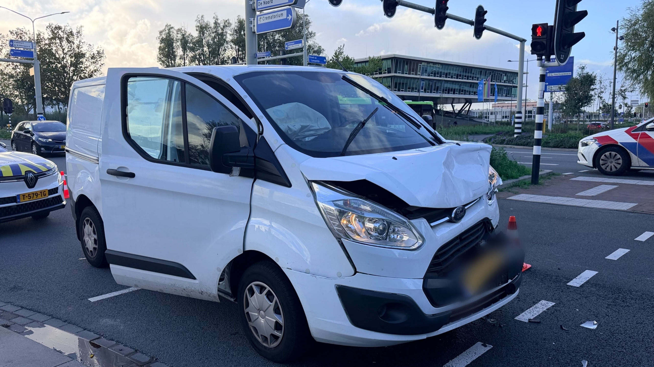 Witte bestelbus met ingedeukte motorkap na aanrijding op kruispunt, met politieauto en pionnen op de weg