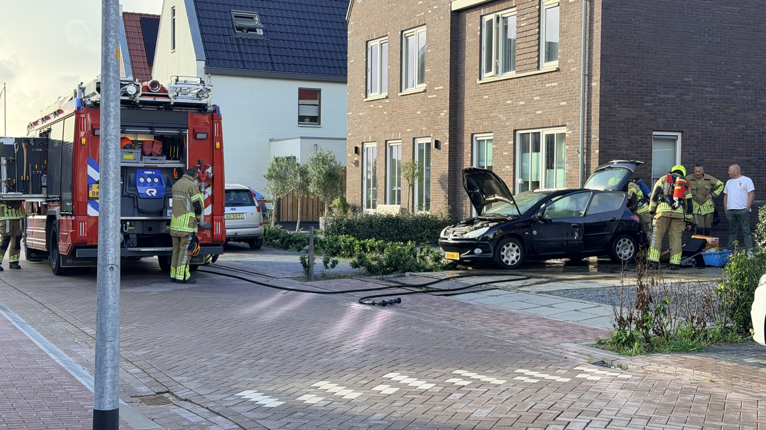 Brandweer bij auto met open motorkap op oprit; brandweerwagen en slangen in woonstraat, omstanders erbij