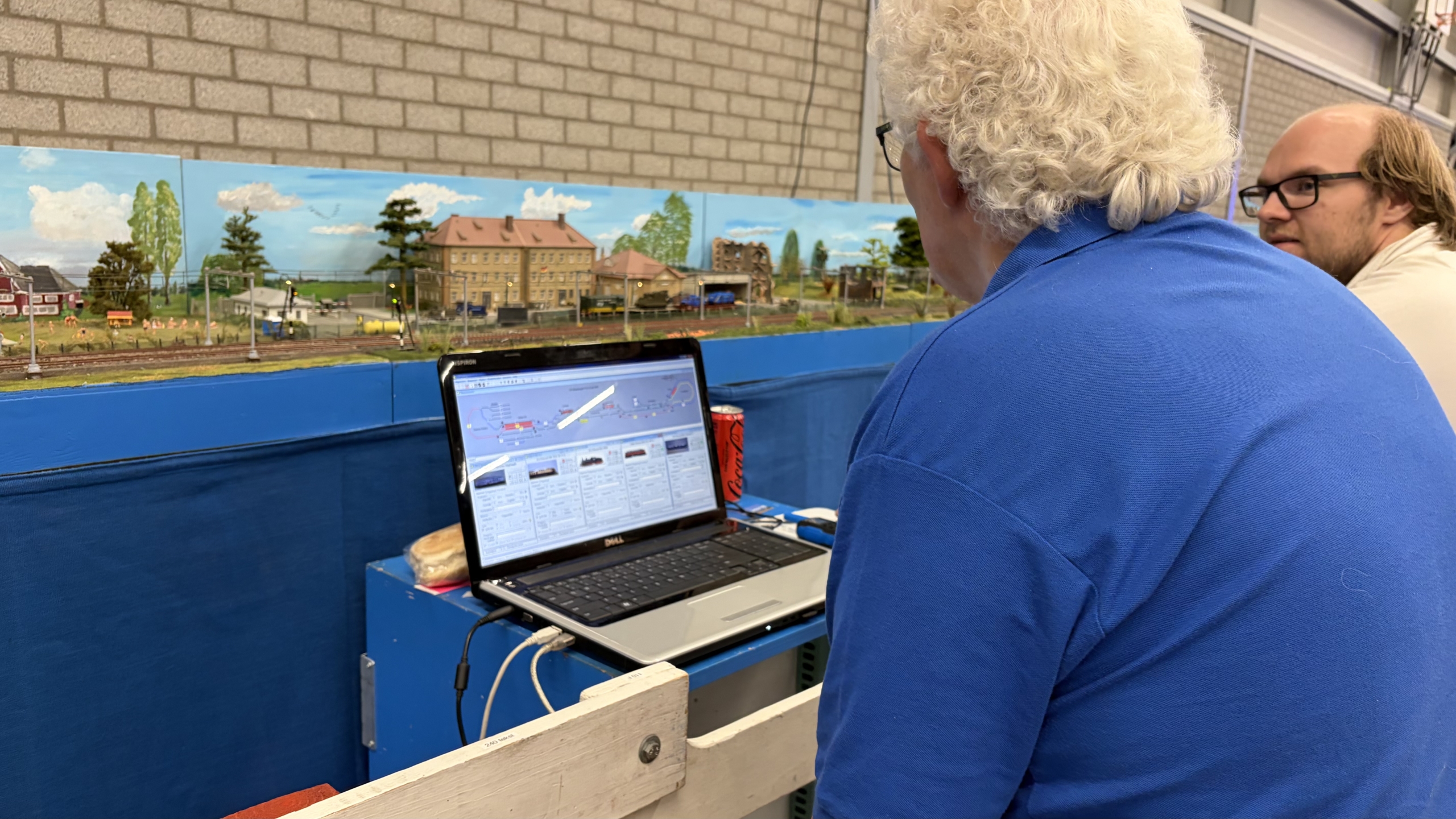 Twee mannen bedienen een modelspoorbaan via een laptop, met op de achtergrond een gedetailleerd miniatuurlandschap