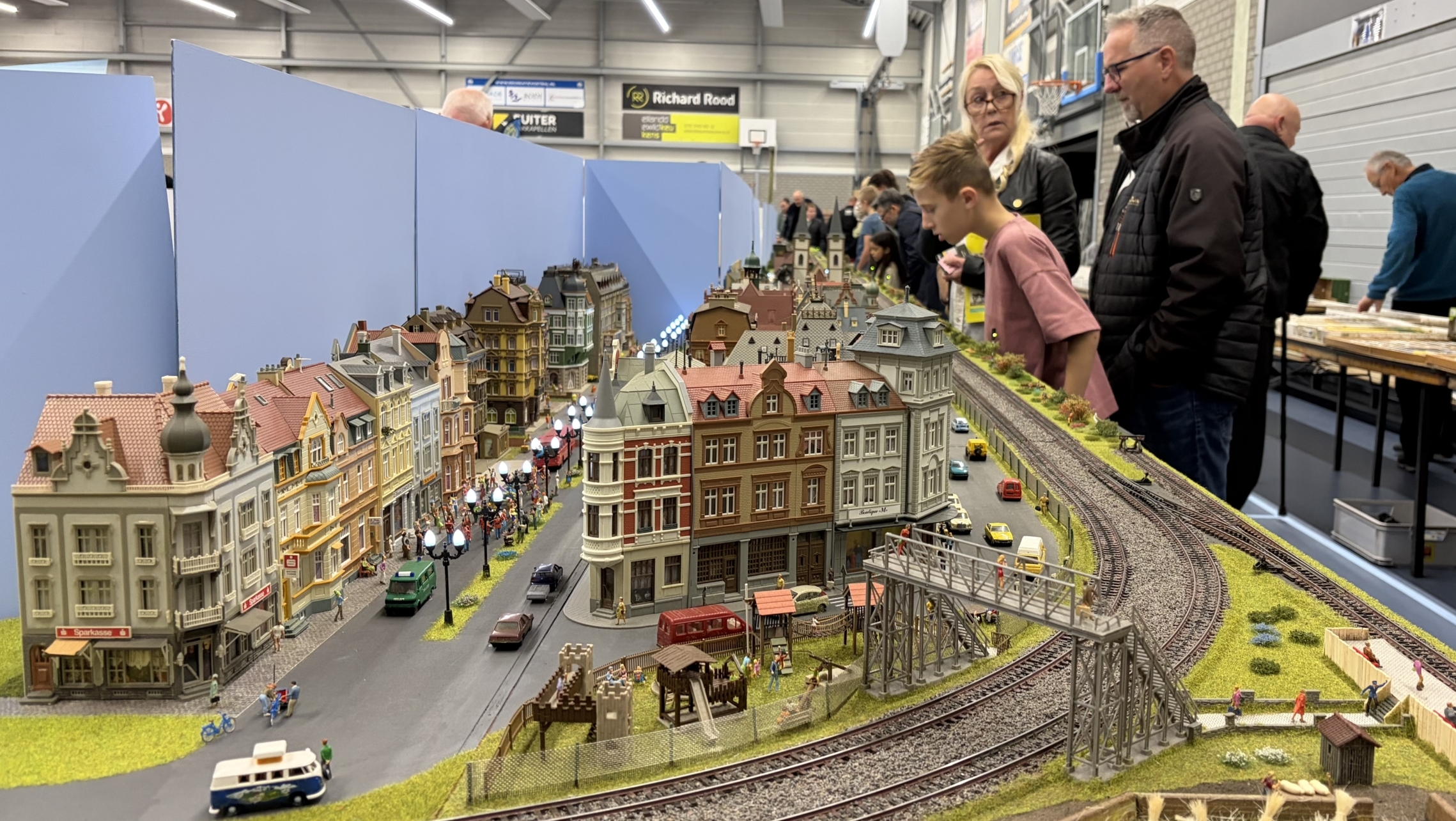 Bezoekers bekijken een gedetailleerde modelspoorbaan met stadshuisjes, straatverlichting en spoorrails in een hal