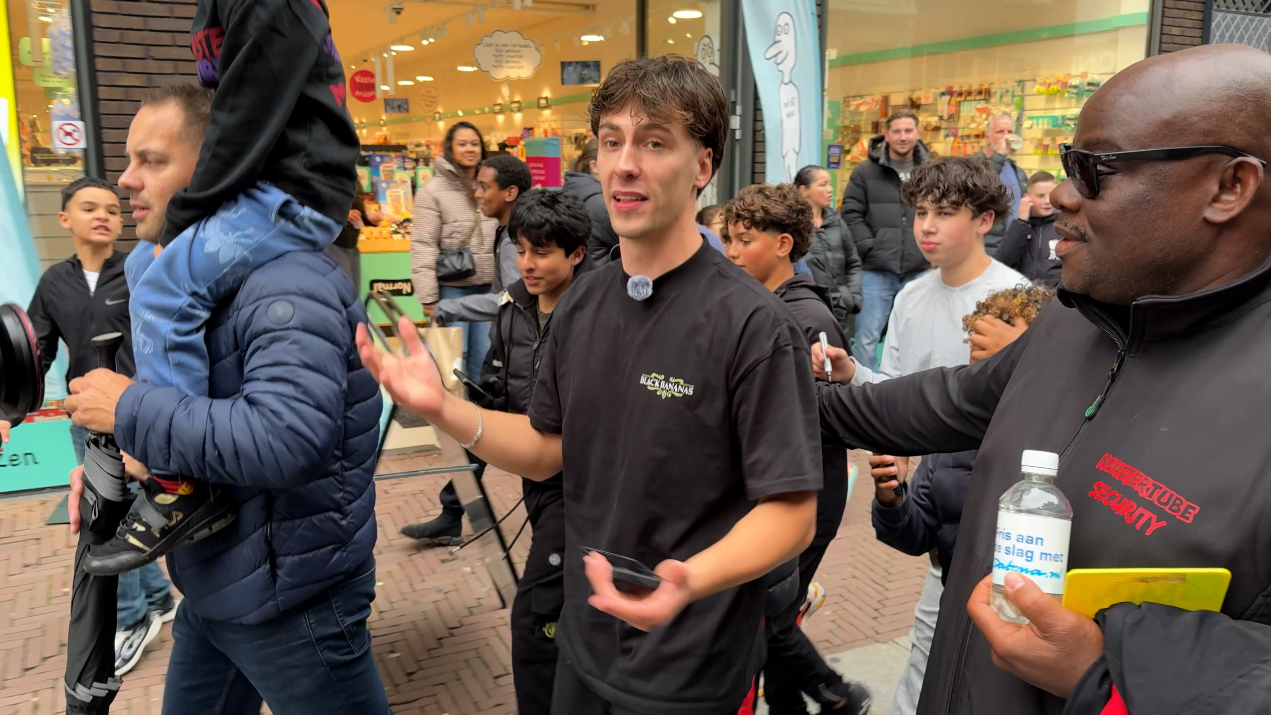 Man met microfoon en telefoon praat al lopend door drukke winkelstraat, naast beveiliger en vader met kind op schouders